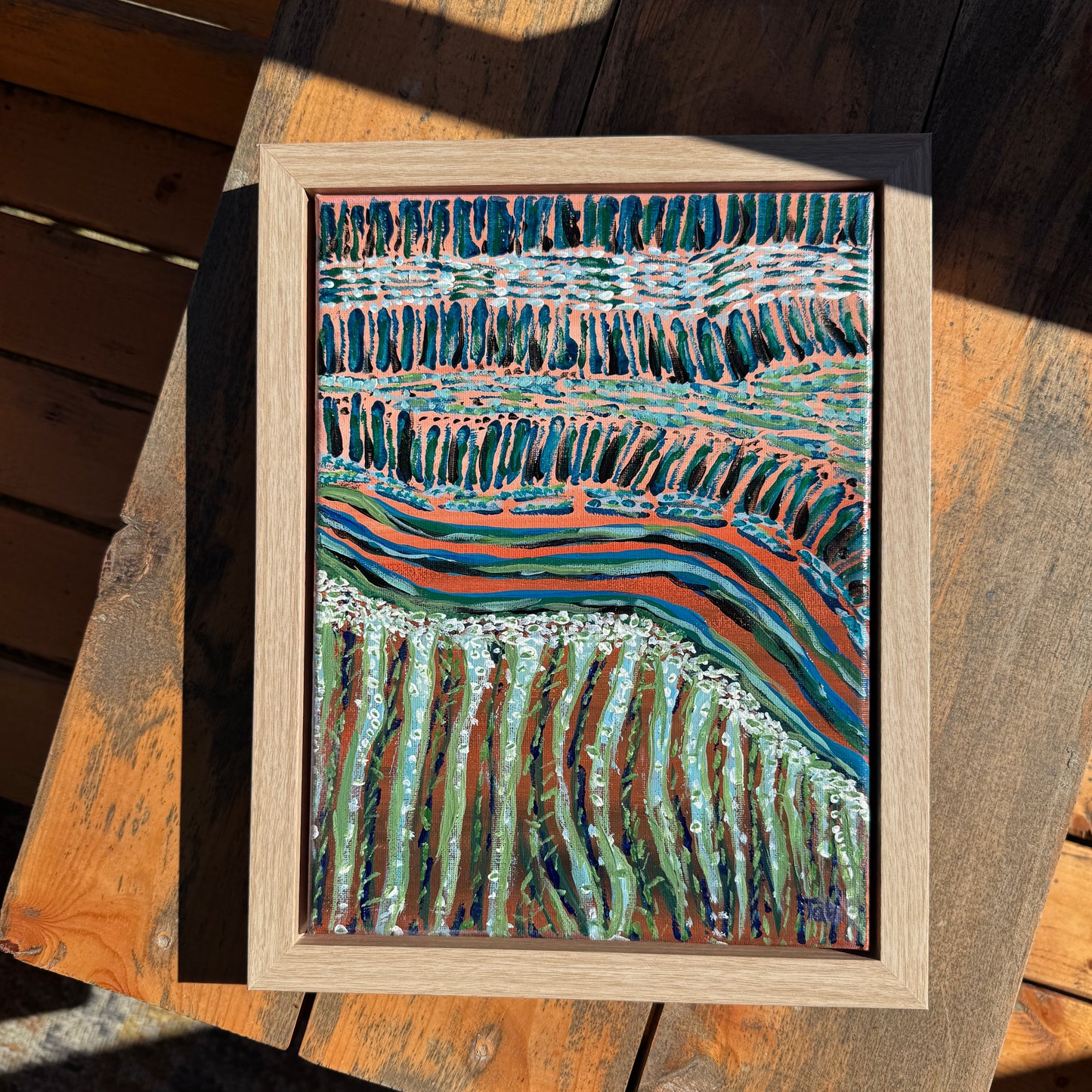 A small abstract pink and green painting on a wooden table, shining in the sunlight.