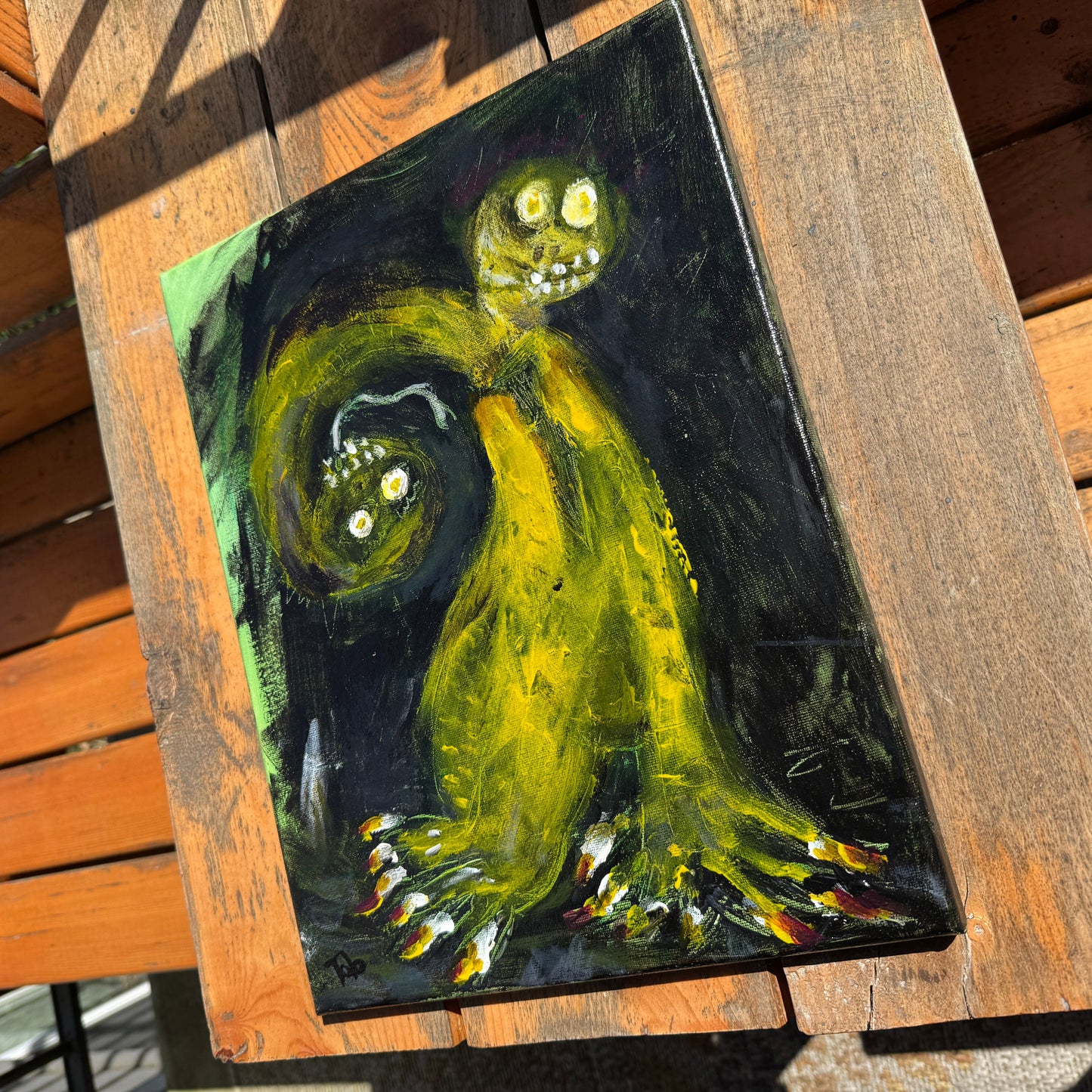Mixed-media artwork, Toxic Boss, on a sunlit wooden table. The strong angle highlights the uncanny abstract art depicting a green two-headed creature with sharp nails on a black background.