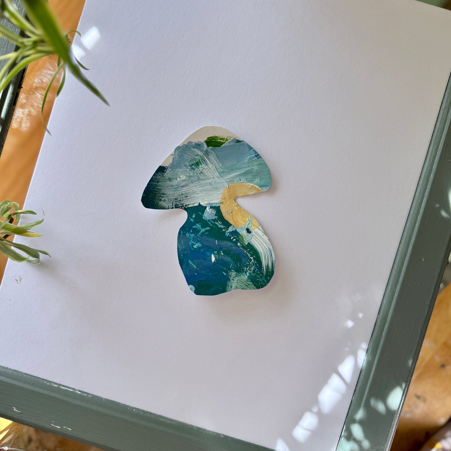 Fun and funky sticker handmade from a blue, green, and tan artist palette. The sticker is placed on a white surface. Colorful green plants and wood surround the mushroom sticker.