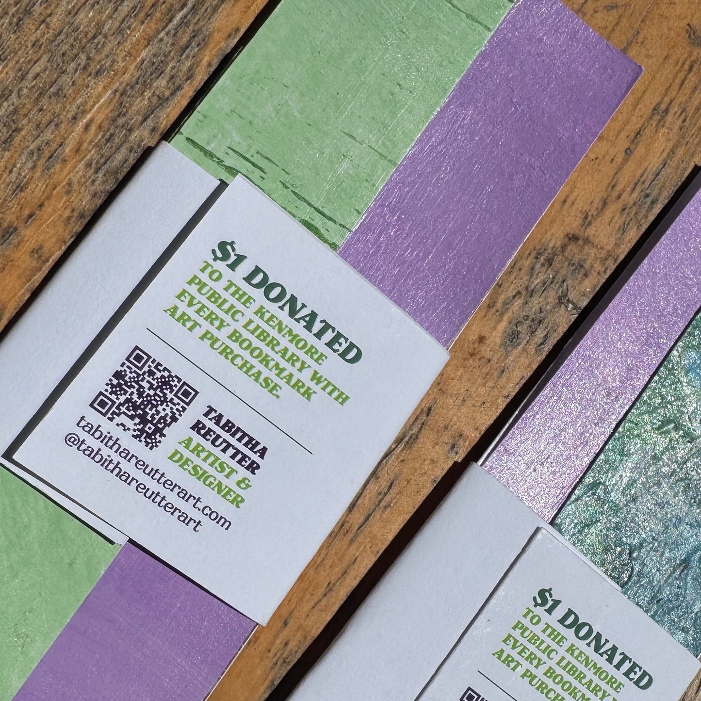 Colorful art bookmarks with a QR code that scans to the artist's website and text on a wooden surface.