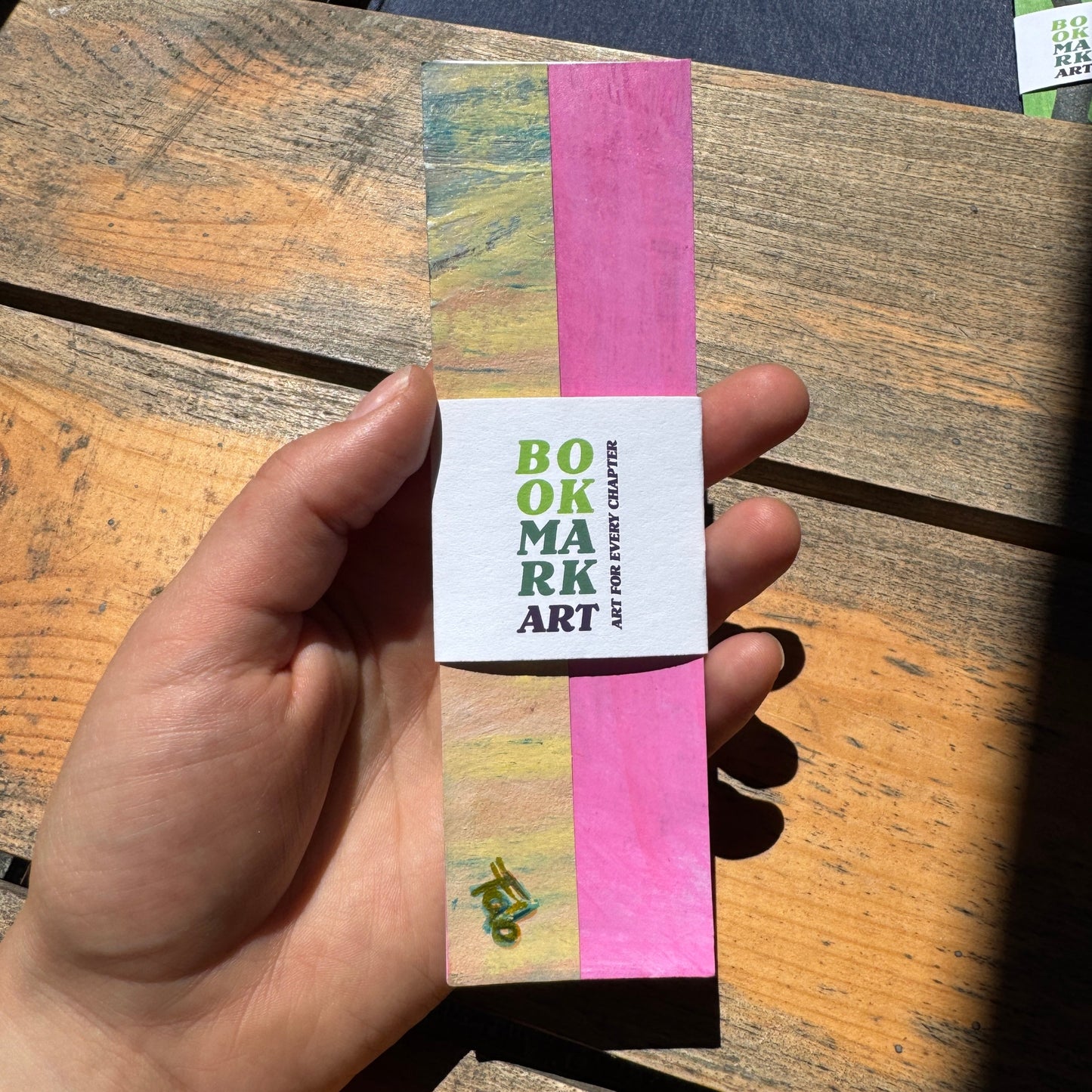 Hand holding a colorful bookmark with 'Bookmark Art' text on a wooden surface.