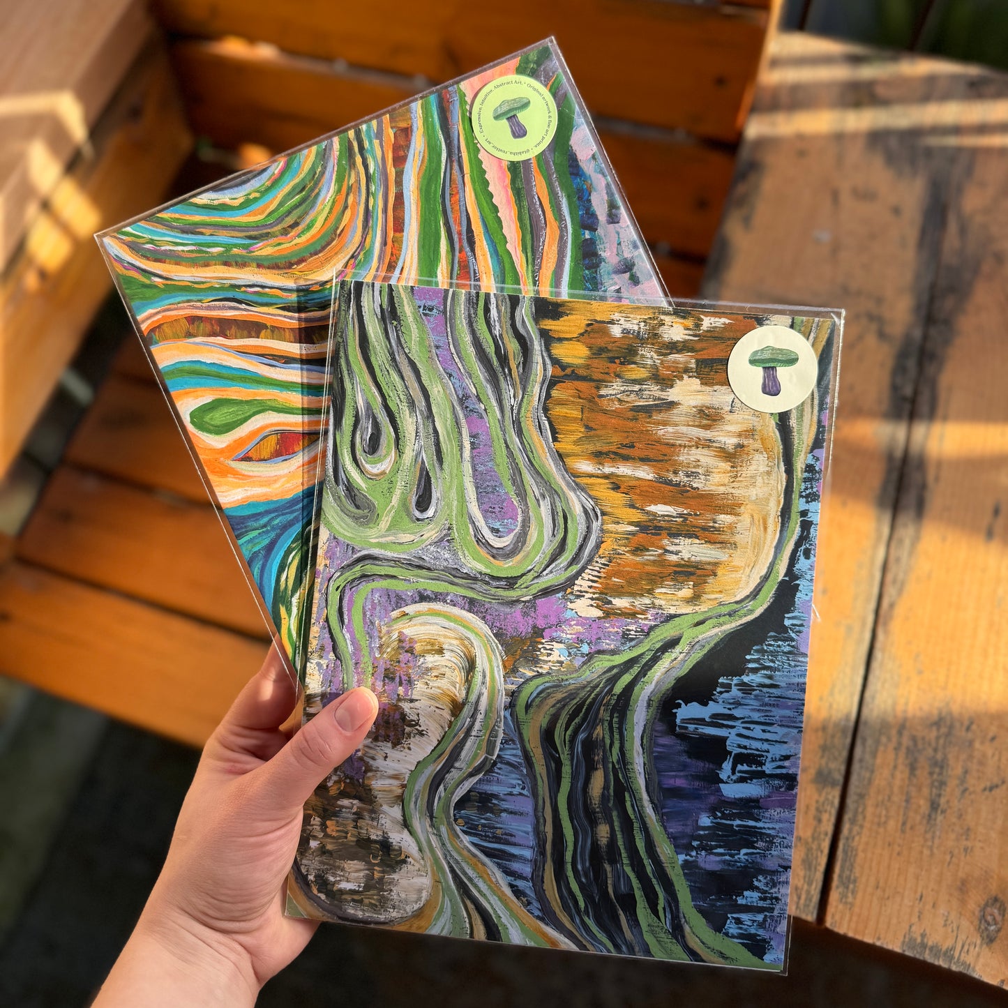 Artist holds two abstract prints, Flowing Through Earth and The Never-Ending Spiral. Prints are vibrant, colorful, and shining in the natural light.