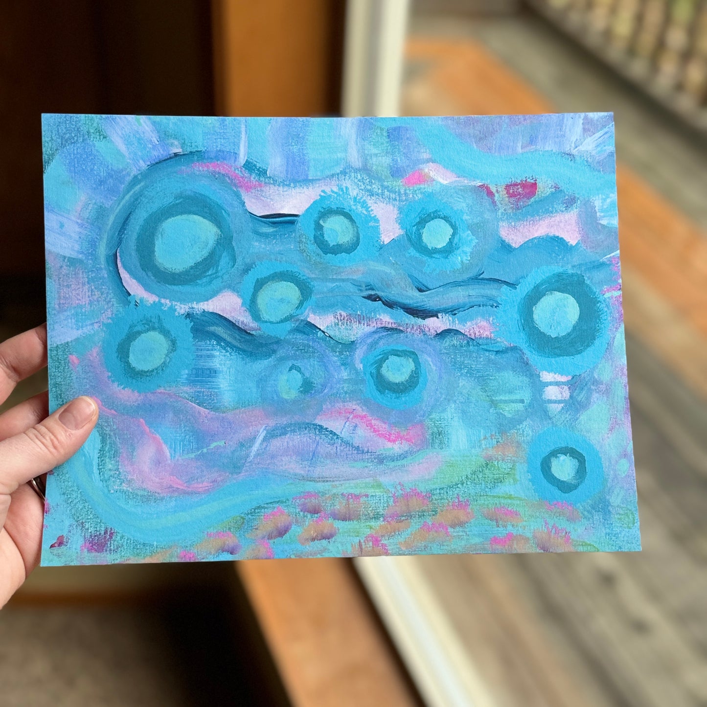 Small, abstract fine art print, Your Weird is Glowing, held by the artist near a window, facing a deck in nature. Abstract artwork features vibrant colors, combining playful design with organic textures.