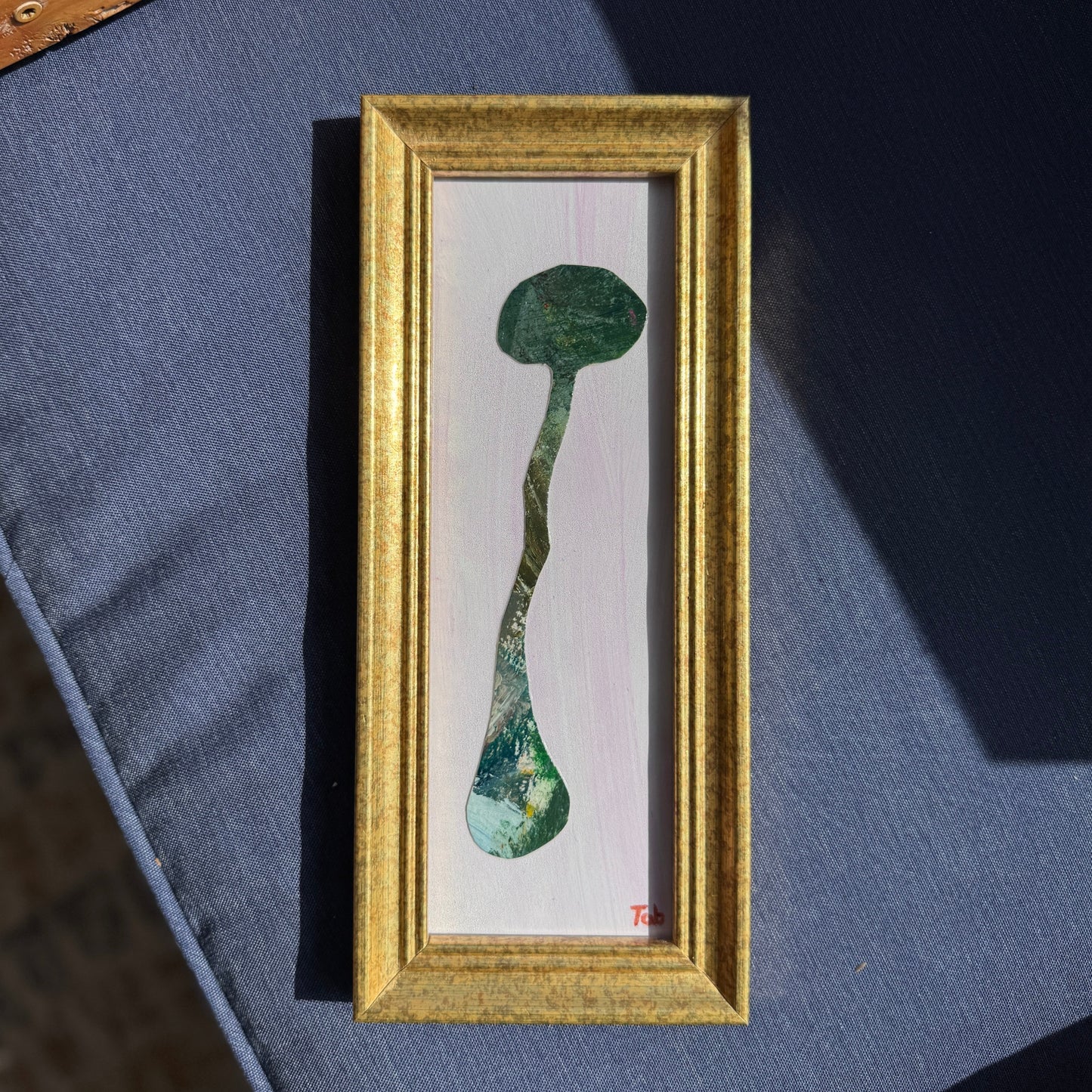 Framed artwork of a long green paper collaged mushroom on a blue surface.