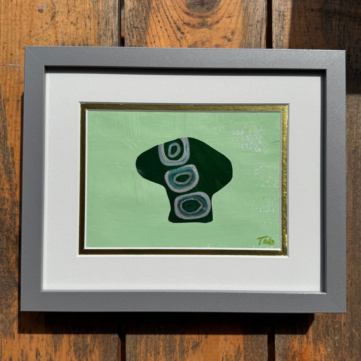 Small green mushroom collage artwork in silver frame on a wood background.