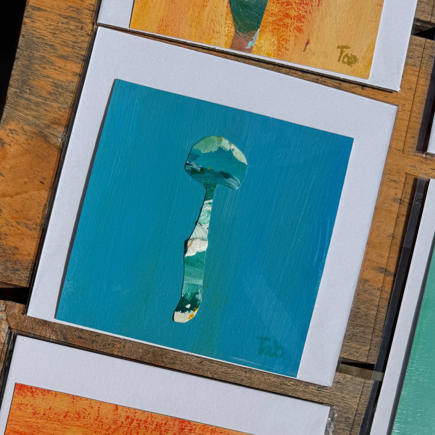 Small blue mushroom collaged artwork with a white backing on a wood surface.