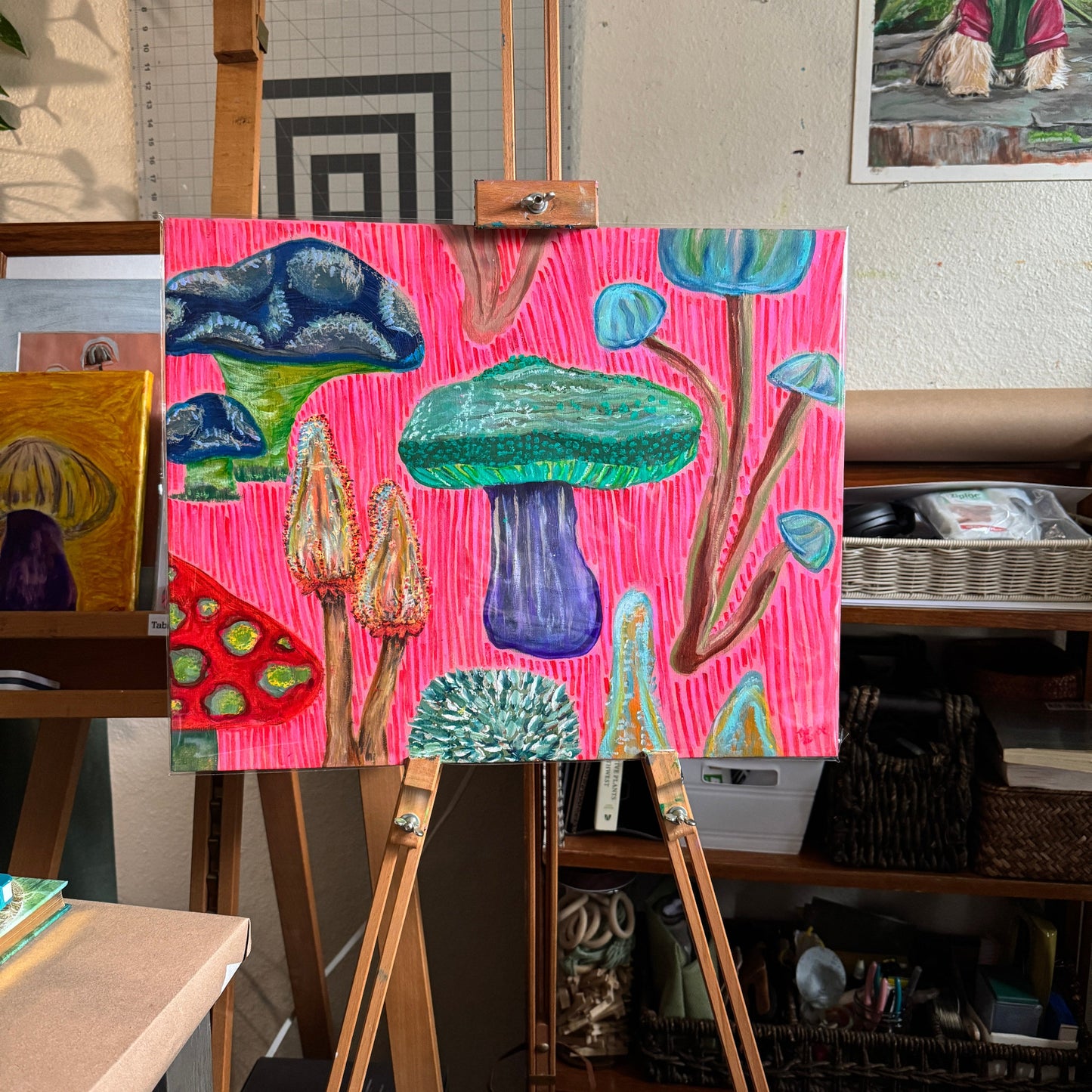 Colorful multimedia artwork Fun Fungi is displayed on an easel in the artist's studio. Bold hot pink background and magenta lines create a graphic look to the art.