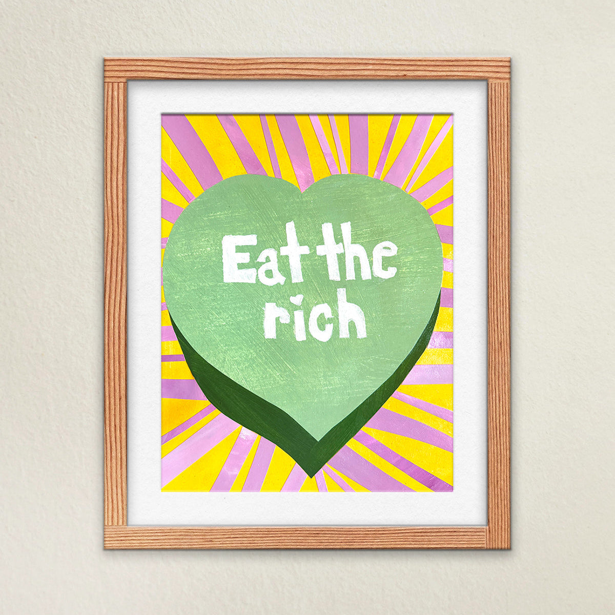Fine art print hanging on a wall in a white matte and wooden frame. The artwork is bright, colorful, and vibrant. Stripes of bright pink and yellow surround a green candy heart with the typeography “Eat the Rich” in the middle.
