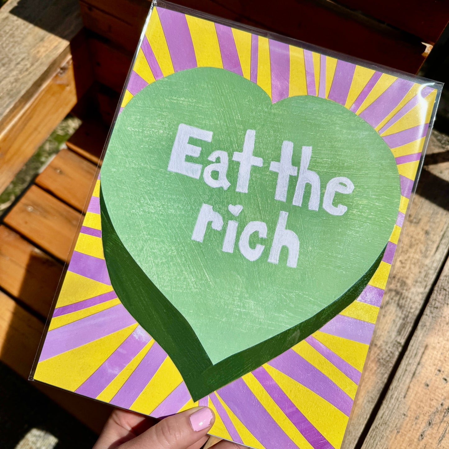 Fine art print held over a wooden bench. The hand holding the print has a pink nail polish. The print shows a pastel green heart with the text “Eat the Rich” in the middle, like a Valentine's Day candy heart. Stripes of bright pink and yellow surround the heart.