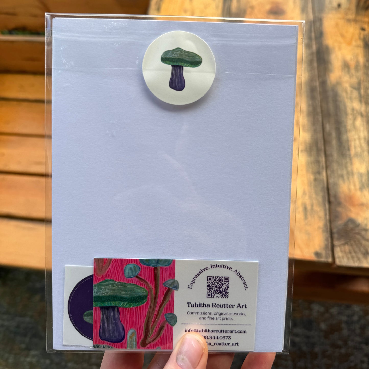 Small, abstract fine art print, held by the artist outdoors by a wooden bench. The back of the print displays a beige sticker with a mushroom and a business card inside the sealed cellophane sleeve to protect the archival inks.