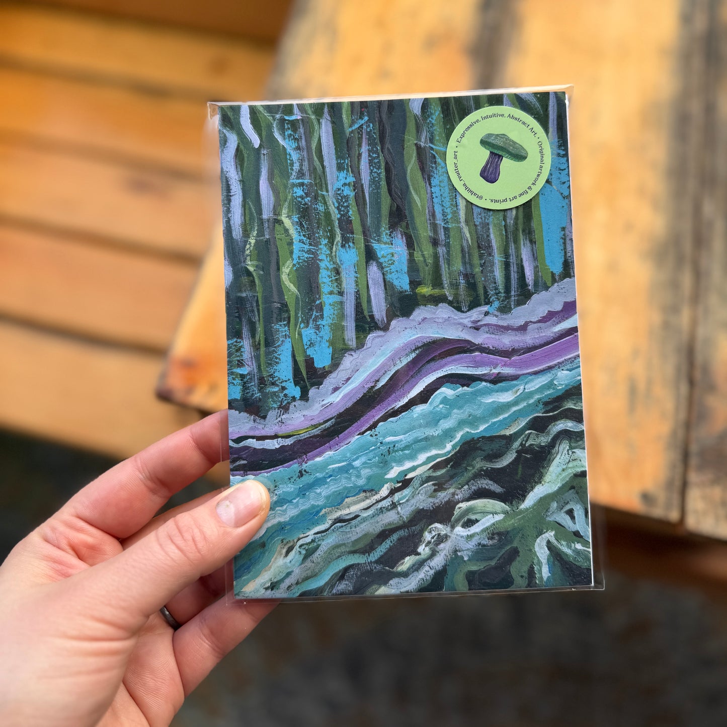 Small, abstract fine art print, By the Water, held by the artist outdoors by a wooden bench. The print has a cellophane wrap and a green sticker with a mushroom.