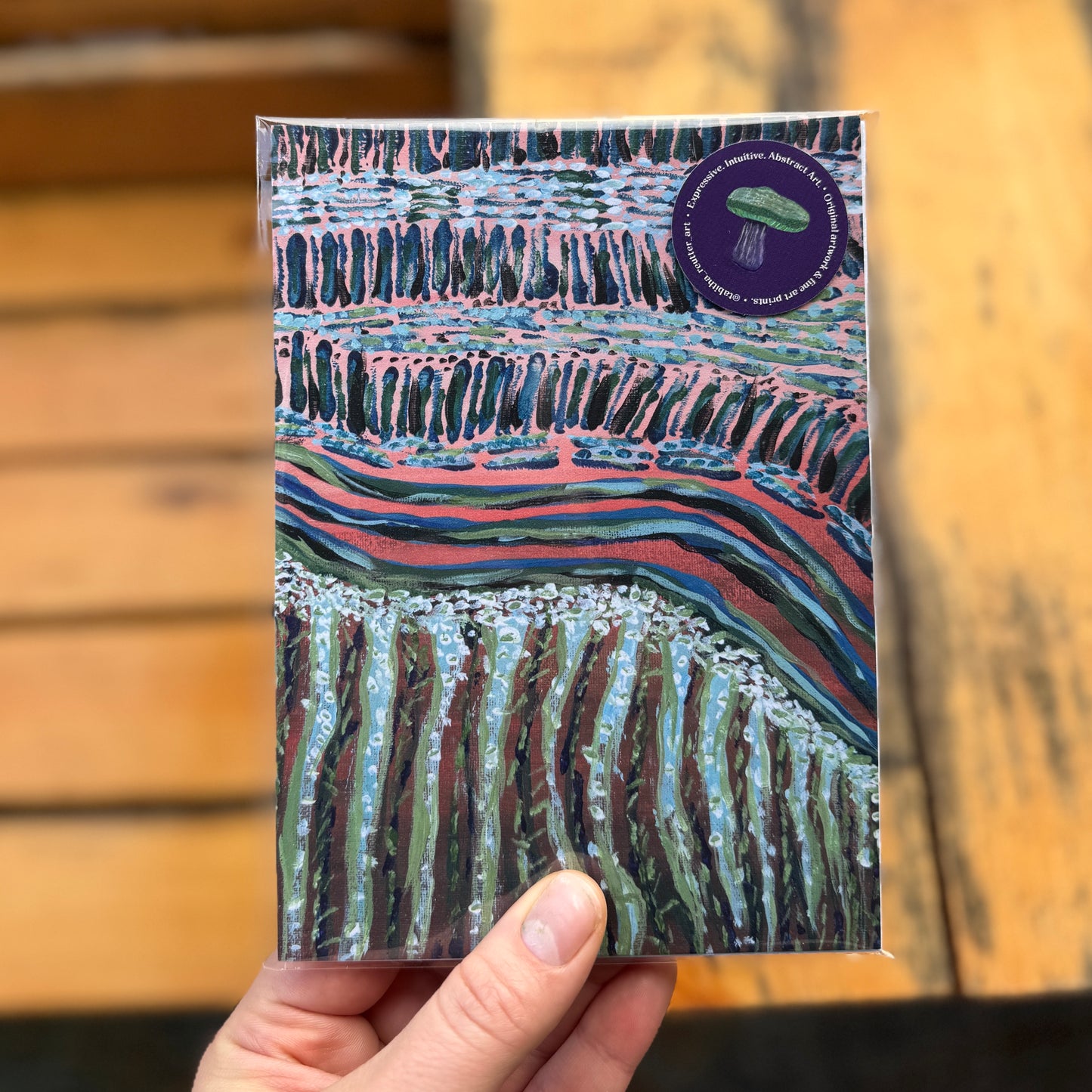 Small, abstract fine art print, Move Along Everyone, held by the artist outdoors by a wooden bench. The print has a cellophane wrap and a purple sticker with a mushroom.
