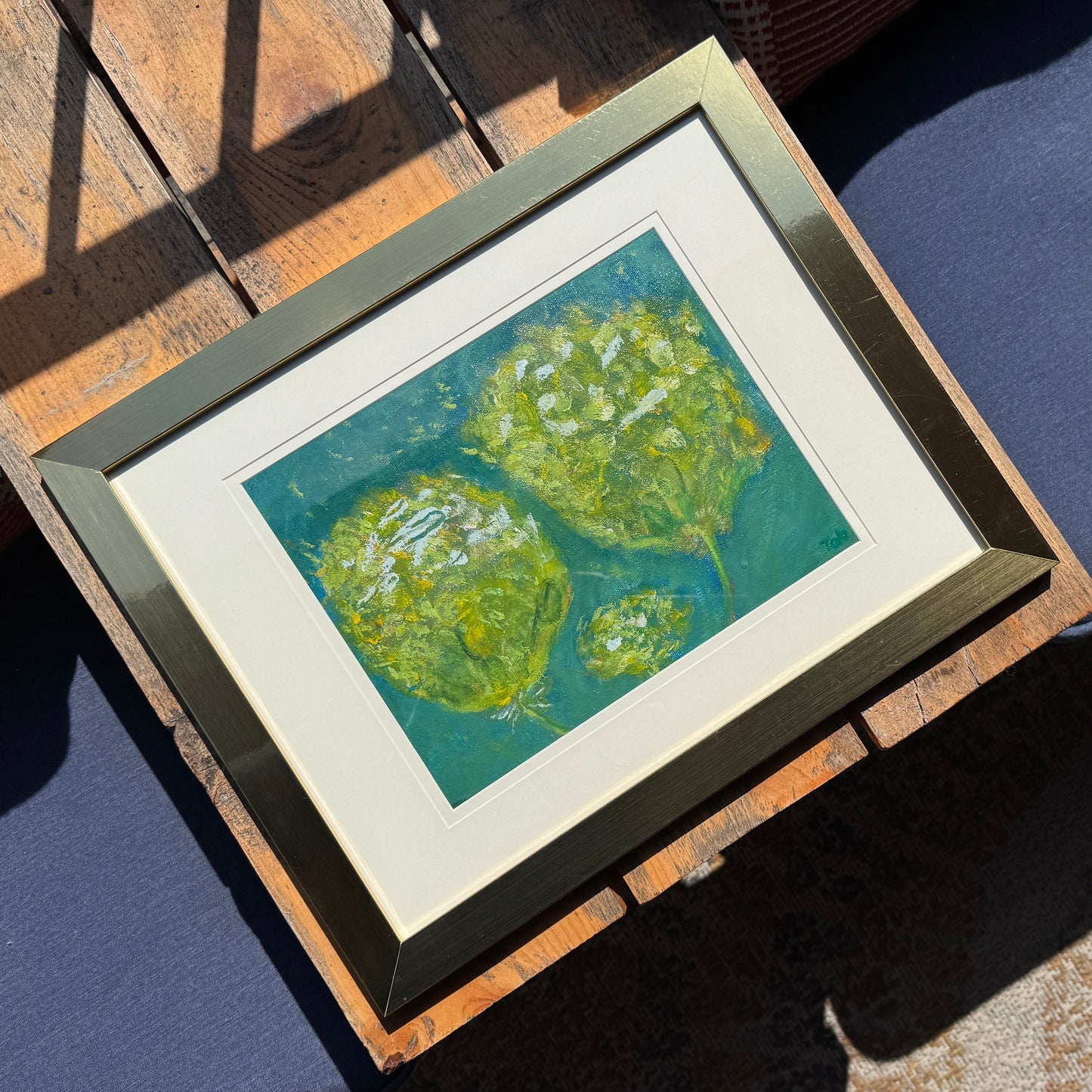 Framed artwork of green abstract fluffy flowers in a golden frame on a wooden bench.