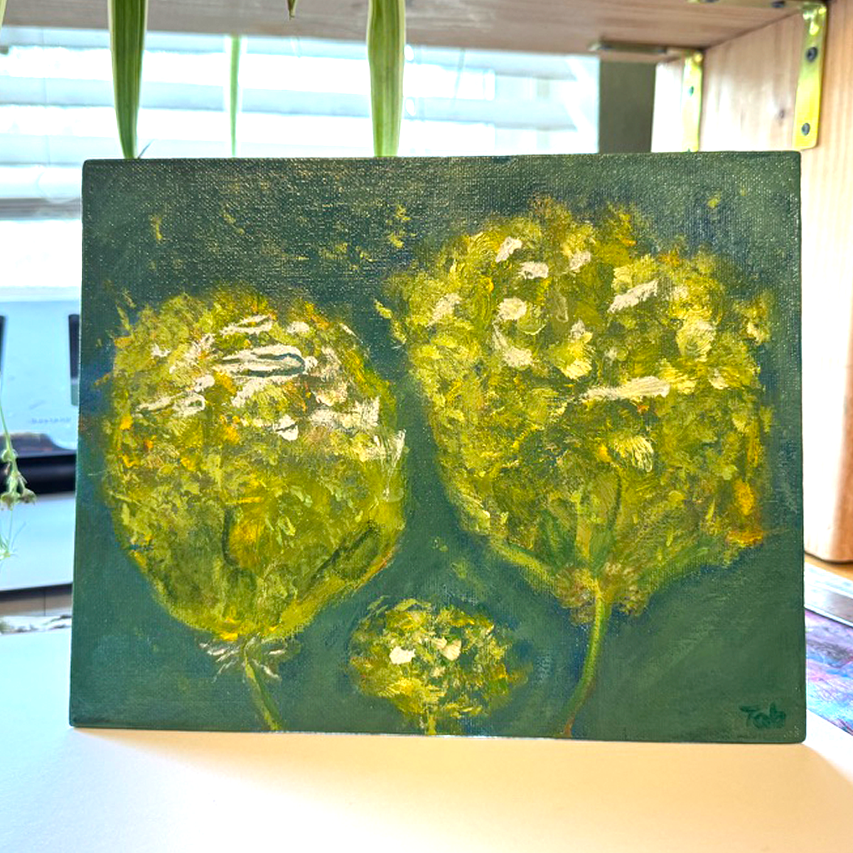 Abstract mixed media painting titled Togetherness, featuring two large yellow flowers with a smaller flower nestled between them. The petals appear to drift and float through the air, set against a painterly green and blue background. The 8x10 canvas artwork evokes connection and harmony with vibrant, expressive brushwork. Shown on a desk. Beautiful light shines through the background.