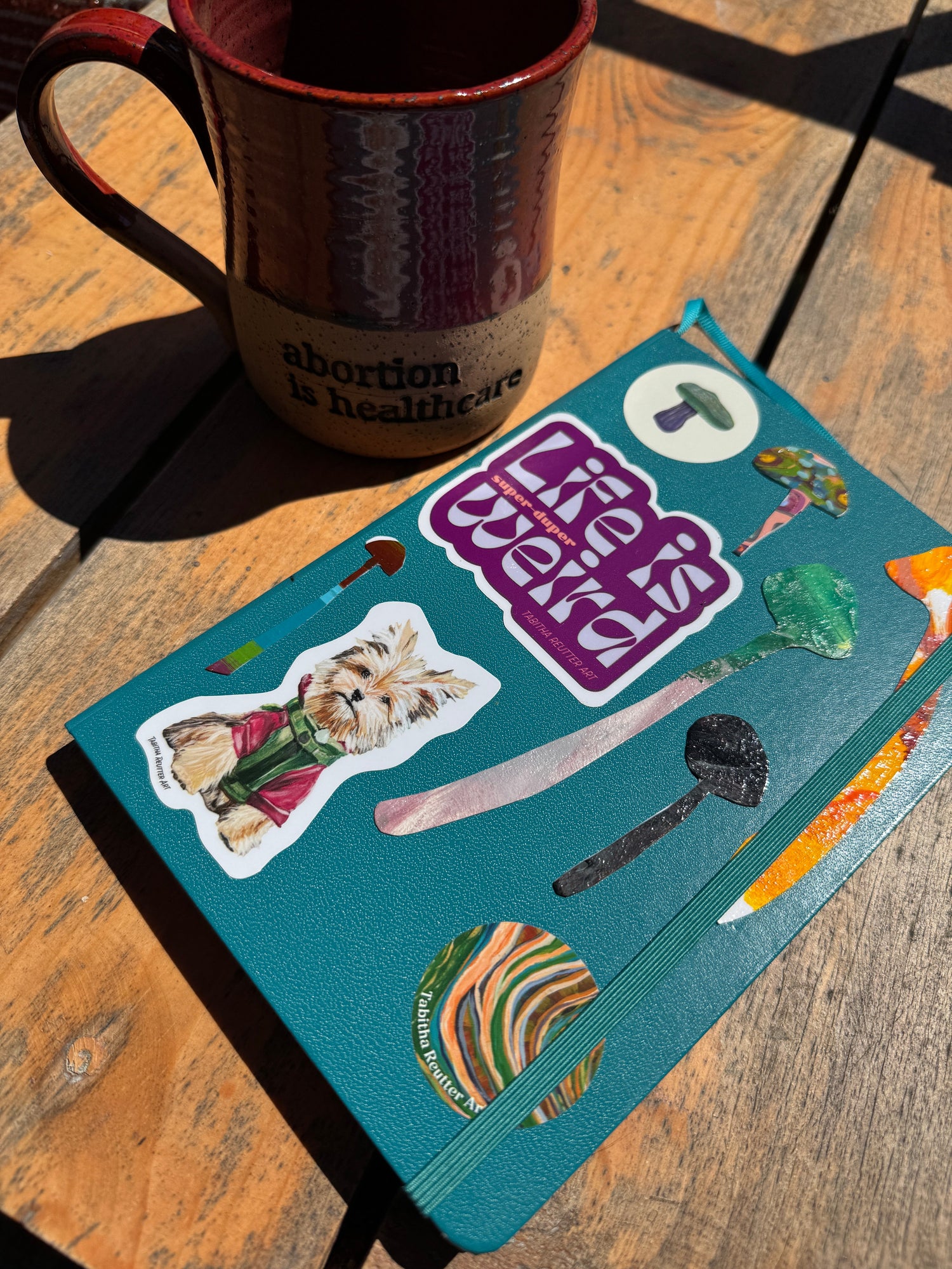 Blue notebook covered in mushroom and vinyl art stickers laying outside on a wooden table next to a rustic maroon ceramic mug