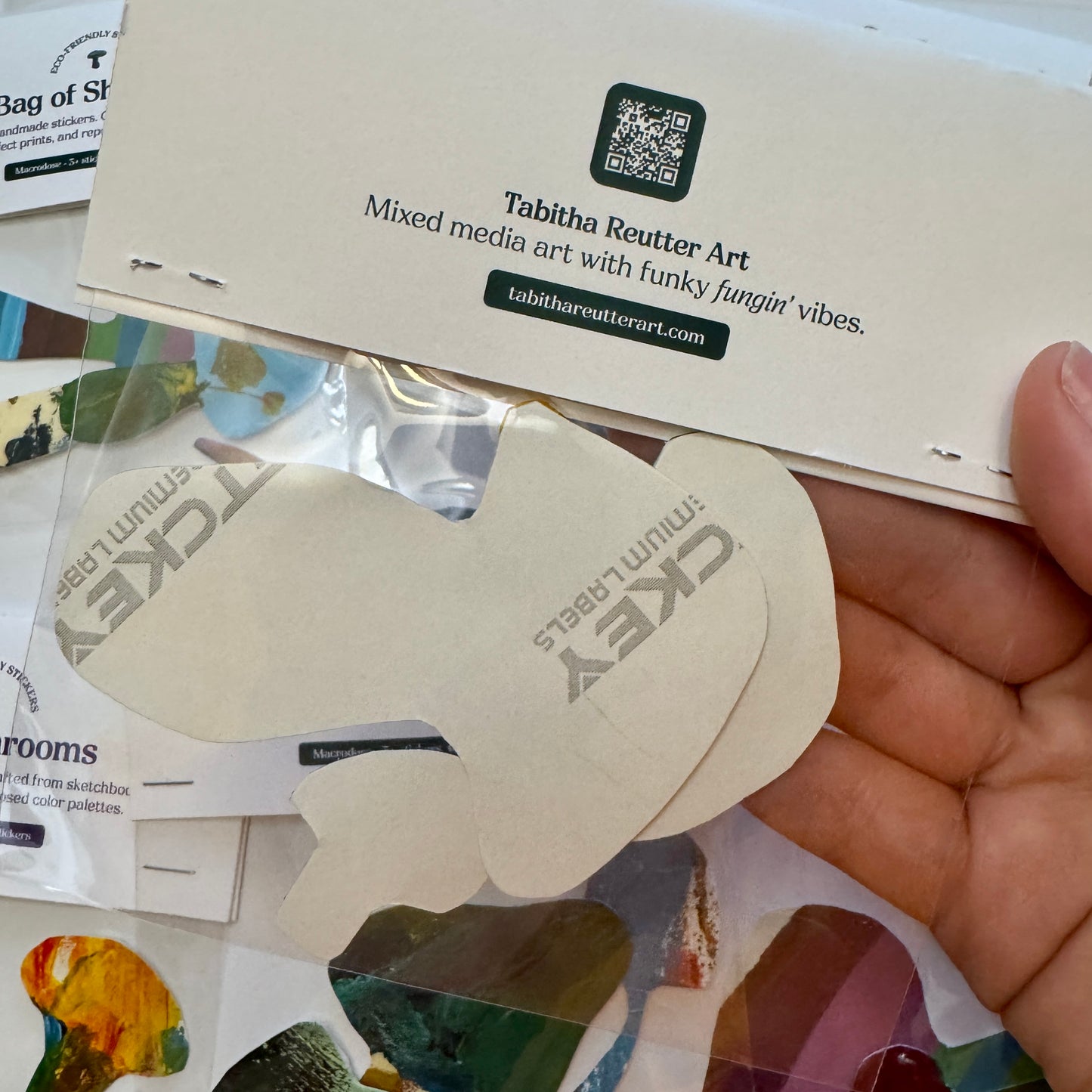 Hand holding a bag of mushroom stickers, showing the back of the stickers. On the packaging at the top, it is white and stapled to the clear cellophane bag that holds the mushroom stickers.