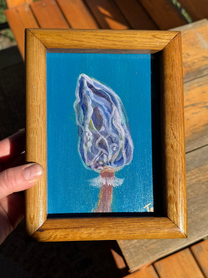 Small blue mushroom artwork held by the artist. Behind the painting, a wooden bench in the sunlight is shown.