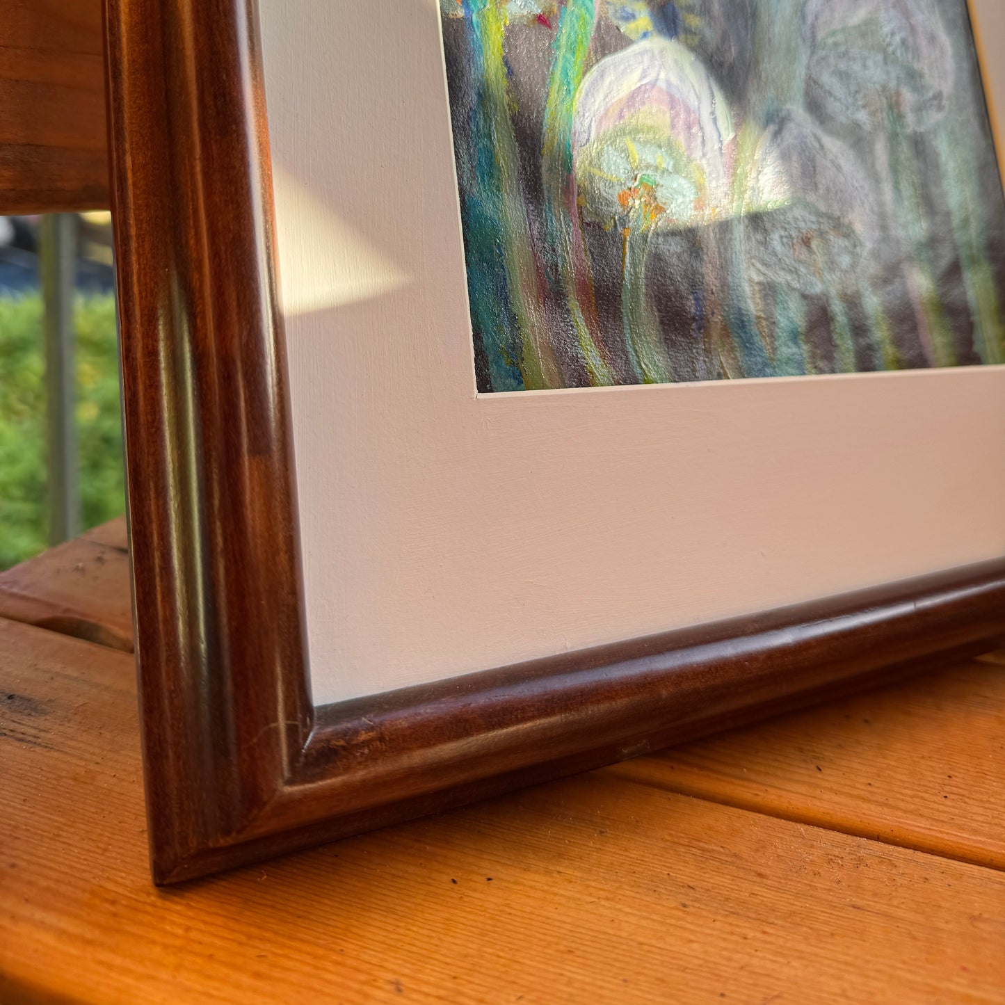 Abstract nature artwork, Mingling Mush featuring purple and green mushrooms with a white matte, in a thrifted, refurbished frame. The artwork is on a wooden table outdoors on a sunlit day. This detail shows the corner of the thrifted frame