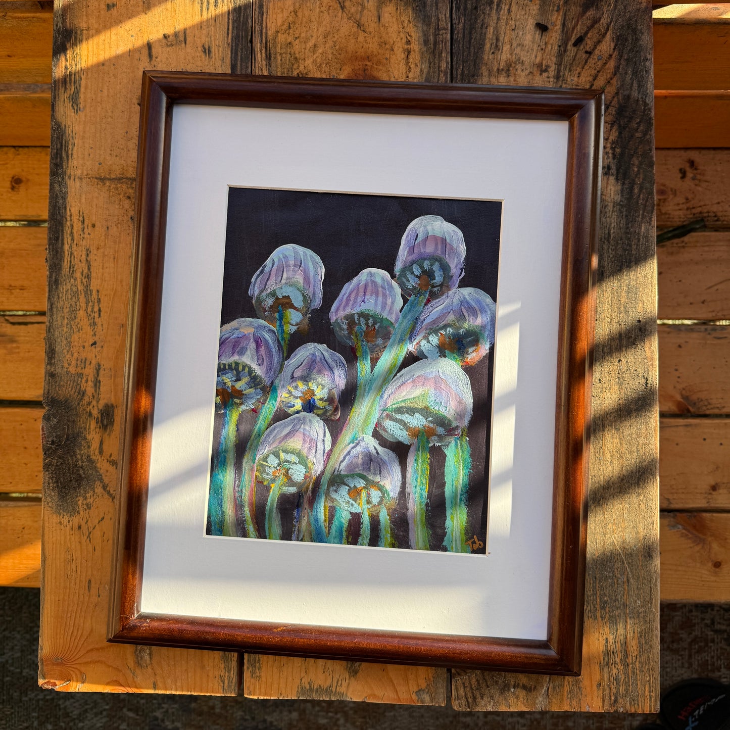 Abstract nature artwork, Mingling Mush featuring purple and green mushrooms with a white matte and a dark walnut refurbished, thrifted frame. The artwork is on a wooden table outdoors on a sunlit day.