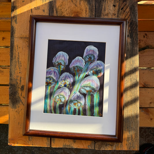 Abstract nature artwork, Mingling Mush featuring purple and green mushrooms with a white matte and a dark walnut refurbished, thrifted frame. The artwork is on a wooden table outdoors on a sunlit day.