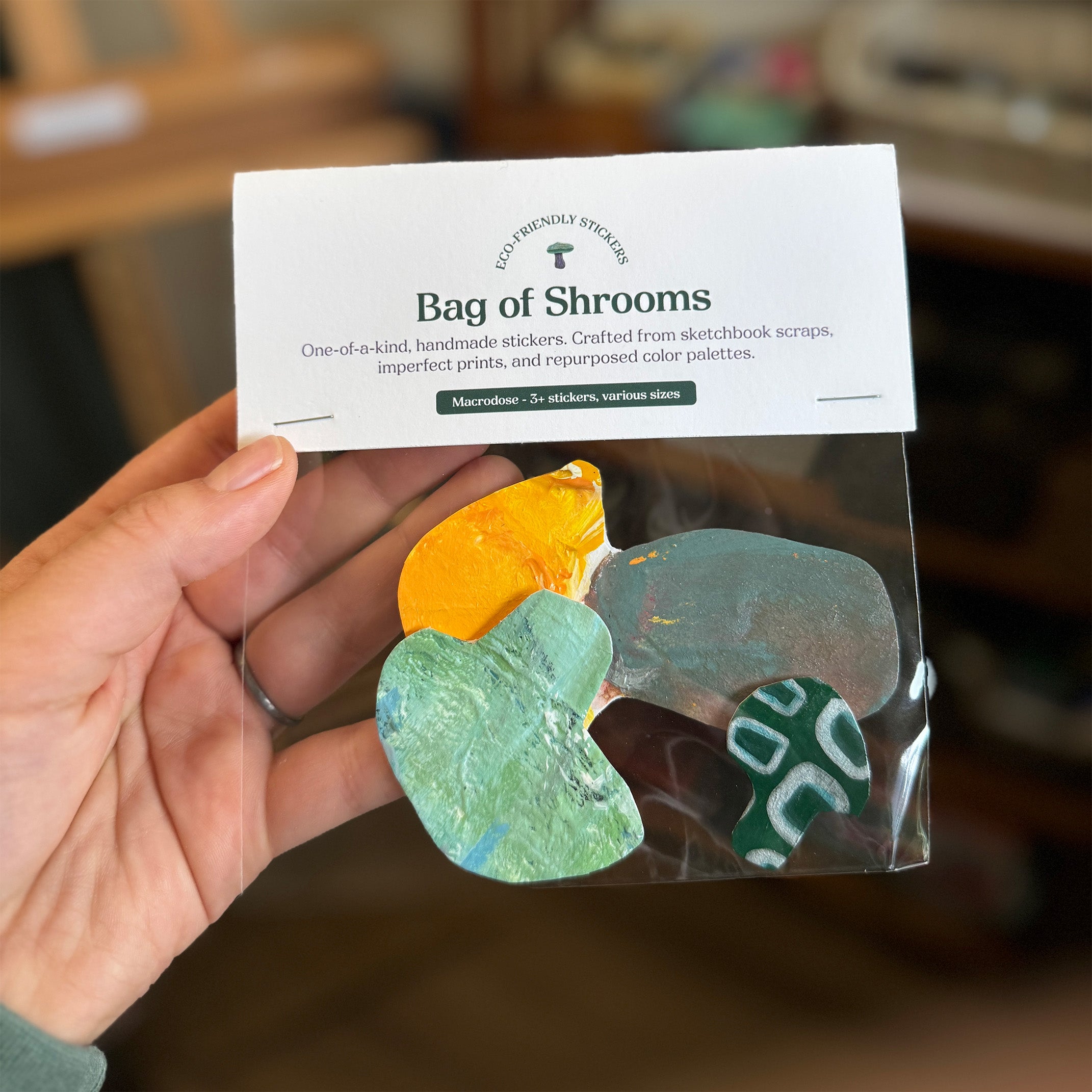 Bag of crafter sticker mushrooms held by a hand. Made from leftover color color palettes. There is a large mushroom with yellow on the tip and gray stem, a green shiny mushroom of medium size, and a small mushroom with a rectangle outline pattern. 