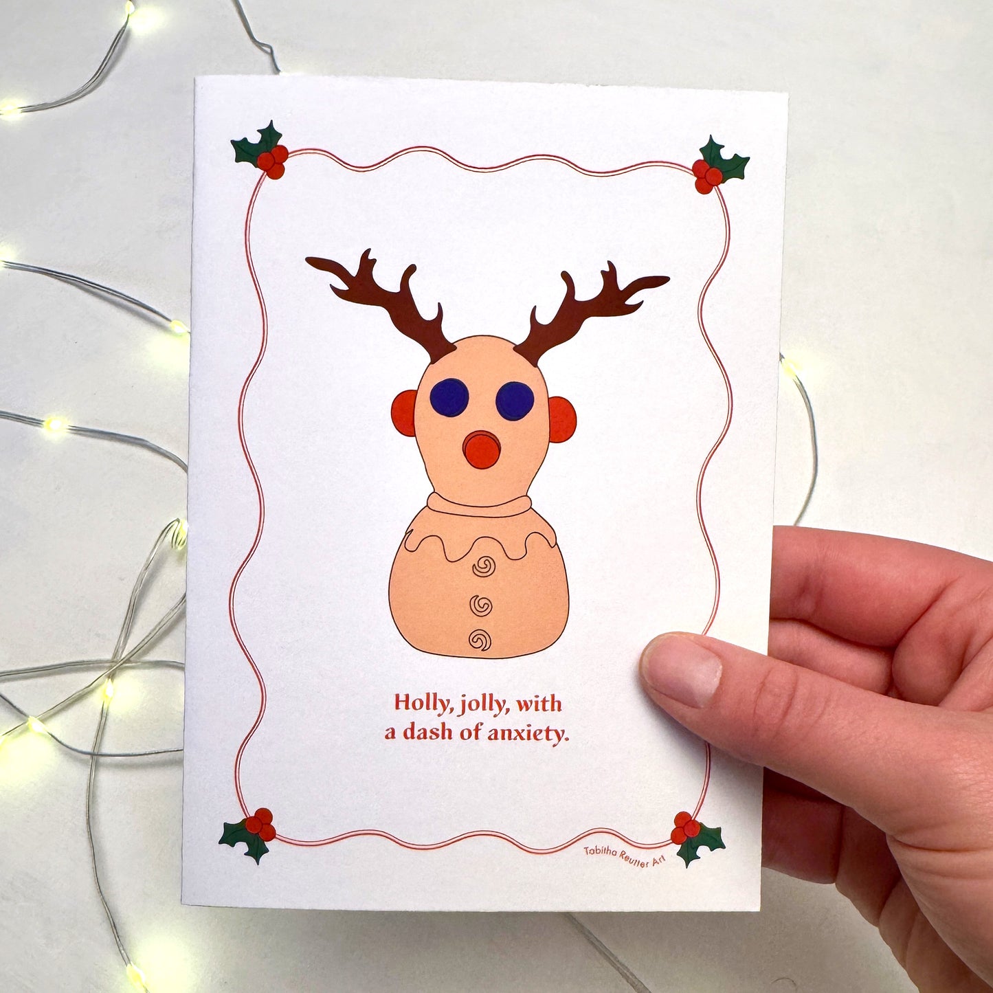 A hand holds a festive Christmas Holiday card featuring a simple graphic illustration of the neurodivergent toy, Panic Pete. Christmas lights flicker in the background on top of a white puzzle board. Holly is illustrated on the card around the image with red wiggly lines.