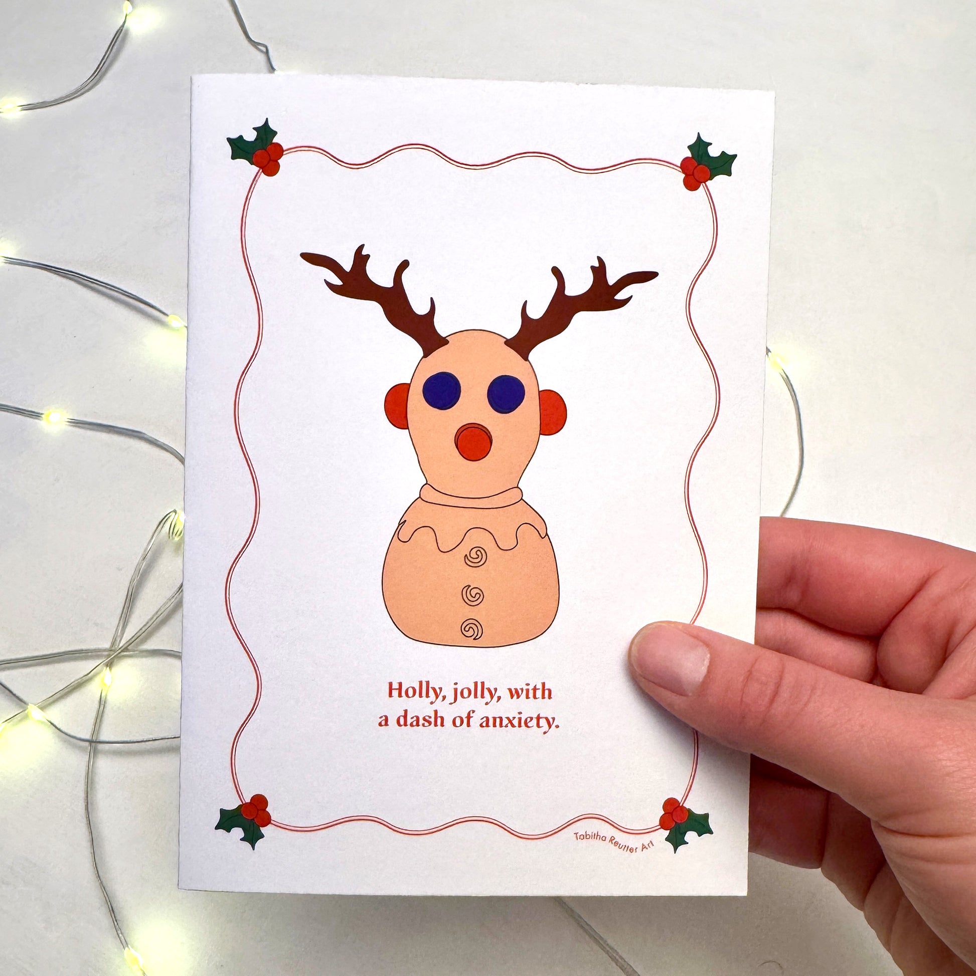 A hand holds a festive Christmas Holiday card featuring a simple graphic illustration of the neurodivergent toy, Panic Pete. Christmas lights flicker in the background on top of a white puzzle board. Holly is illustrated on the card around the image with red wiggly lines.
