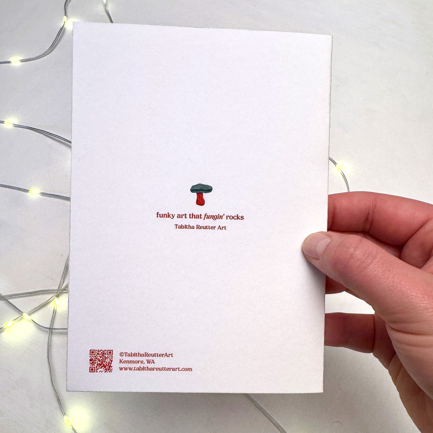 An artist hand holds a Christmas card featuring the back of the white card. a festive green and red mushroom logo with the text 'funky art that fungin rocks — Tabitha Reutter Art‘ in red. Small Christmas lights flicker in the background on top of a white painted background.