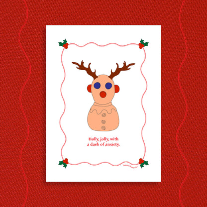 White greeting card displayed on a vibrant Holiday red felt-textured background with an illustration of the squeeze neurodivergent toy, Panic Pete, wearing festive reindeer antlers in the center of the card. Red text, under the artwork, reads: Holly, jolly, with a dash of anxiety.