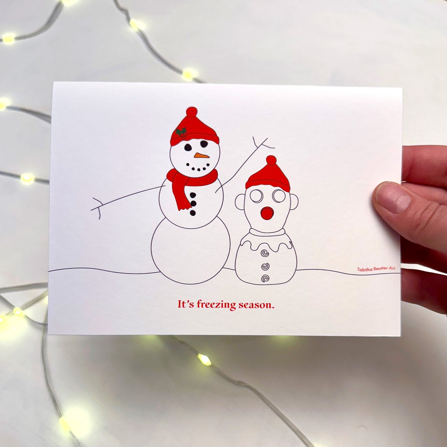 A hand holds a funny Christmas Holiday card featuring a simple graphic line drawing illustration of a snowman next to a snow-person version of the neurospicy squeeze toy, Panic Pete, on a snowy hill. Soft lights flicker in the background on top of a white background.