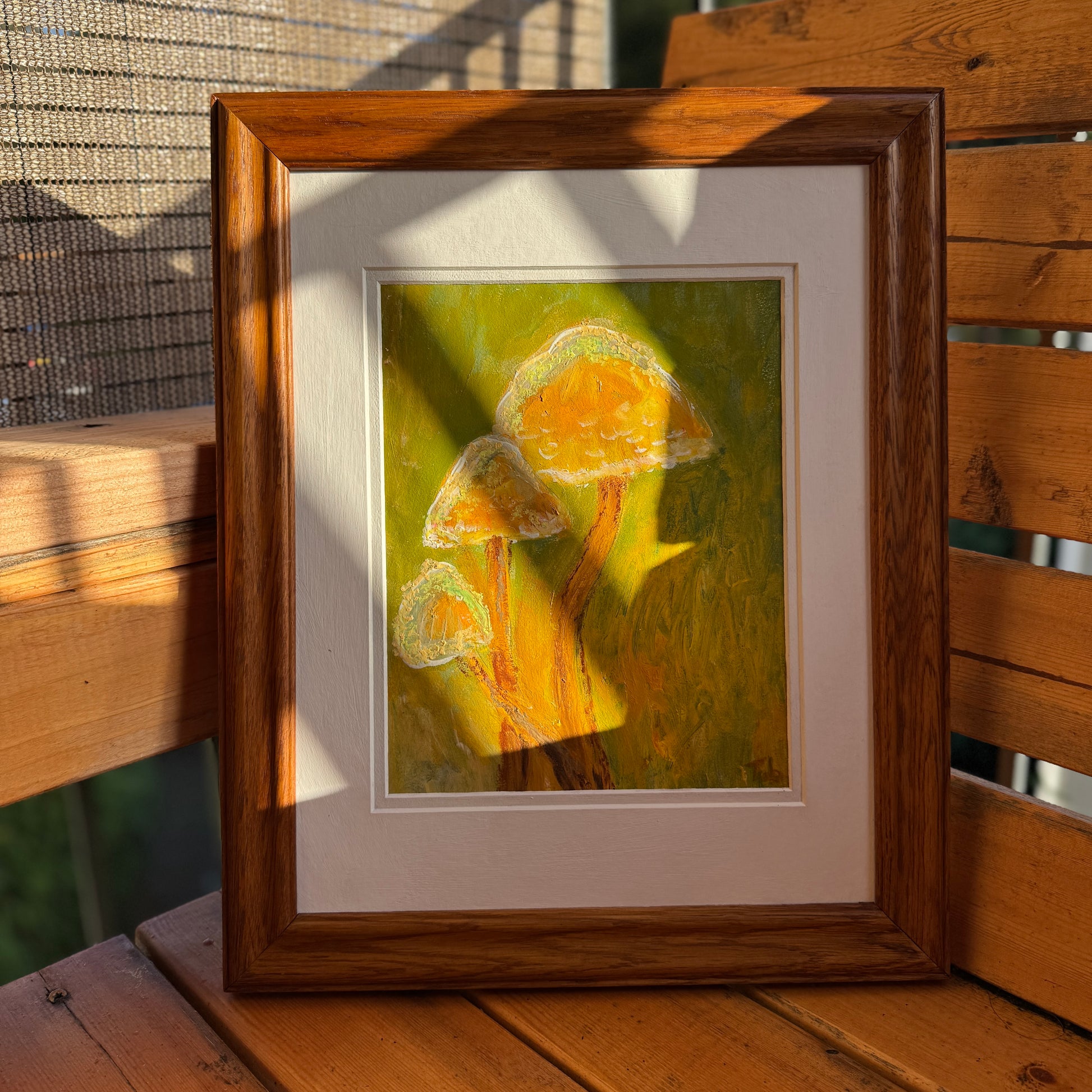 Nature-inspired, multimedia artwork, Golden Hour, on a sunny day on a wooden bench. It is displayed in a thrifted frame with a white matte.