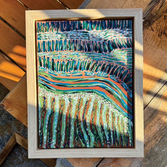 A small abstract pink and green painting lies on a wooden bench in the glistening sunlight. The photographer stands above, their bleached jeans appear at the bottom of the composition along with a wooden frame on the ground.