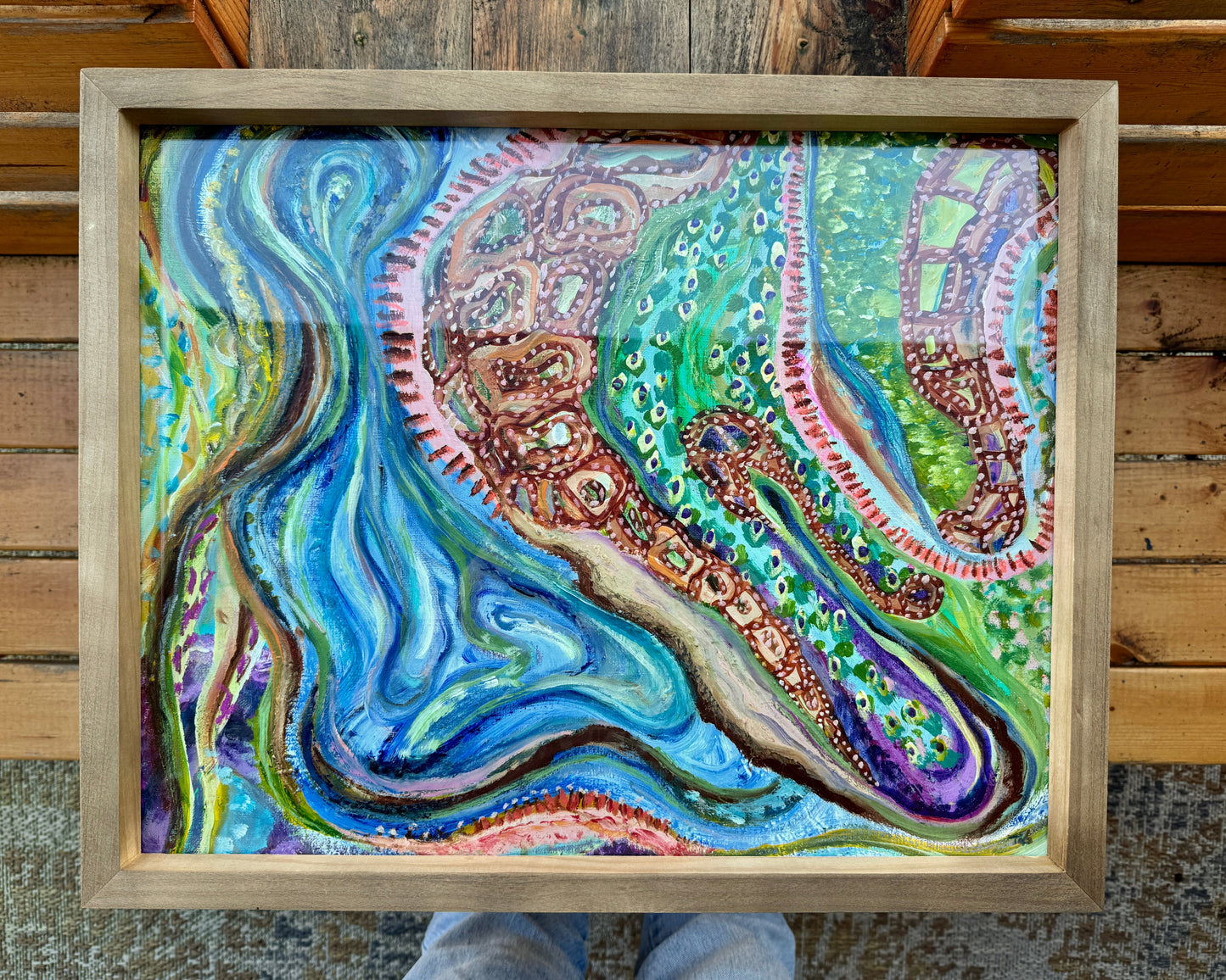 Framed, abstract mixed media artwork, Sitting With Thoughts rests on a wooden bench on the artist’s wooden bench. The frame is a light wood color, and the artwork is bright and vibrant with multicolored hues across colors of blue, green, orange, and yellow.