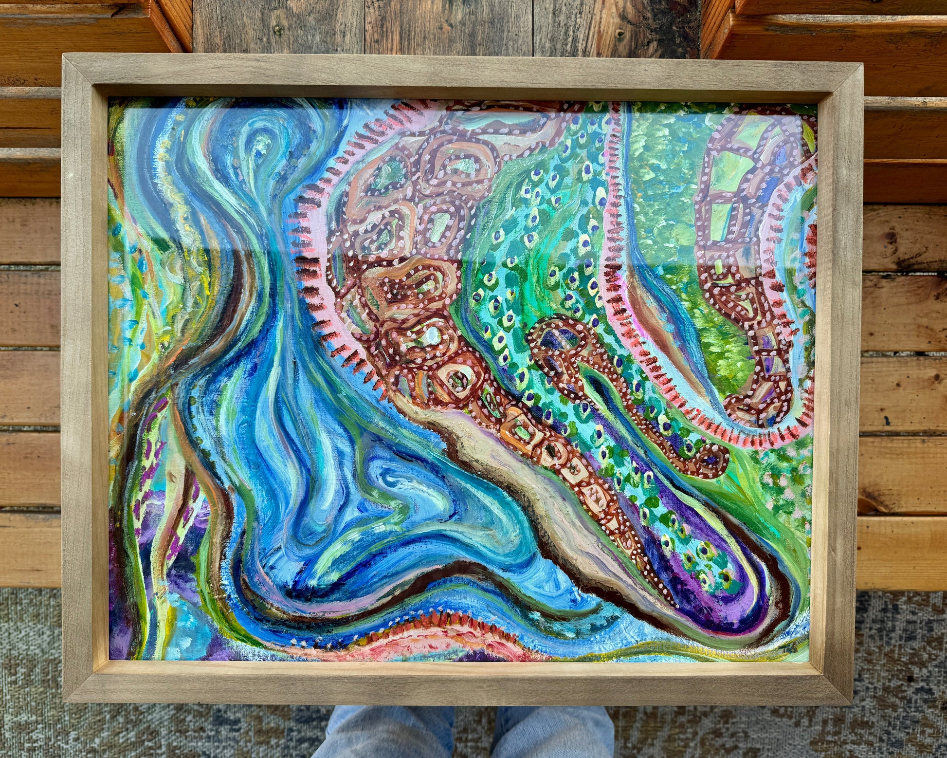 Framed, abstract mixed media artwork, Sitting With Thoughts rests on a wooden bench on the artist’s wooden bench. The frame is a light wood color, and the artwork is bright and vibrant with multicolored hues across colors of blue, green, orange, and yellow.
