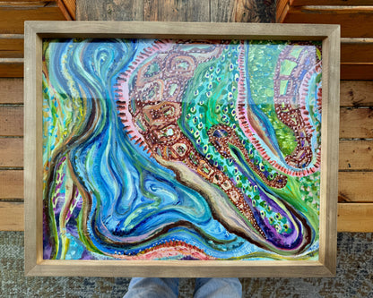 Framed, abstract mixed media artwork, Sitting With Thoughts rests on a wooden bench on the artist’s wooden bench. The frame is a light wood color, and the artwork is bright and vibrant with multicolored hues across colors of blue, green, orange, and yellow.