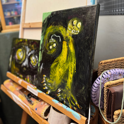 A lifestyle image of mixed-media artwork, Toxic Boss on an artist's easel. The sharp angle shows the abstract art depicting a green two-headed creature with sharp nails on a black background.