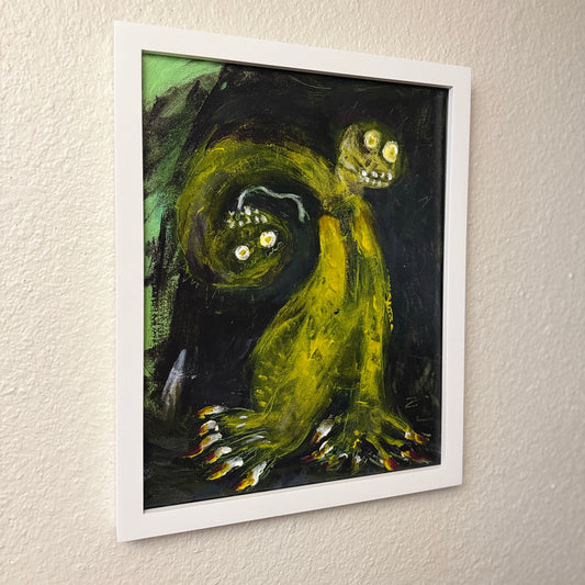 Mixed-media artwork, Toxic Boss, in a white wooden frame hanging on a textured white wall. The abstract artwork features a green two-headed creature with sharp white teeth, bulging eyeballs, and a snake-like tongue.