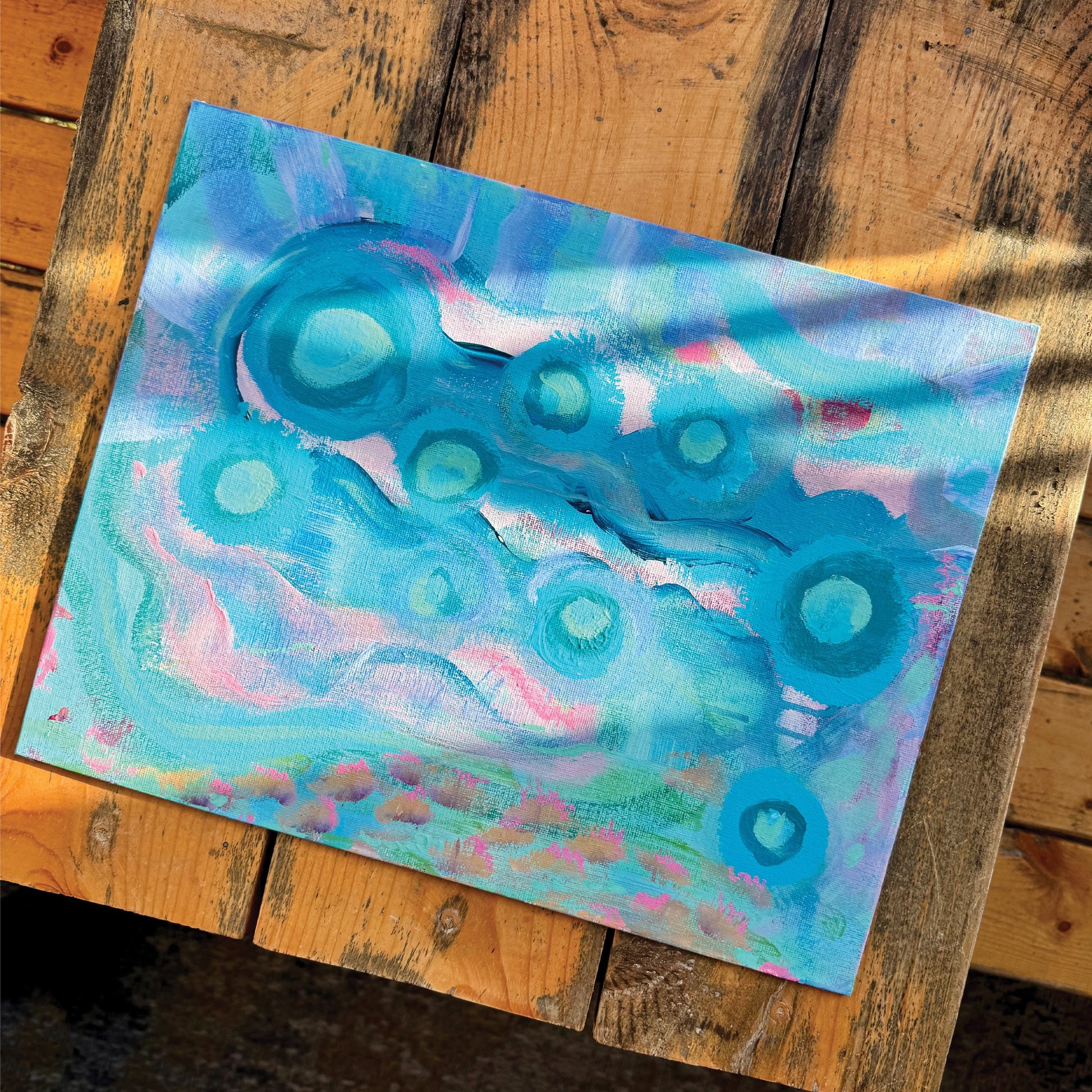 Abstract artwork, Your Weird is Glowing, on a wooden bench in the sunlight. Baby blue and pink colors intermingle, creating a whimsical effect.
