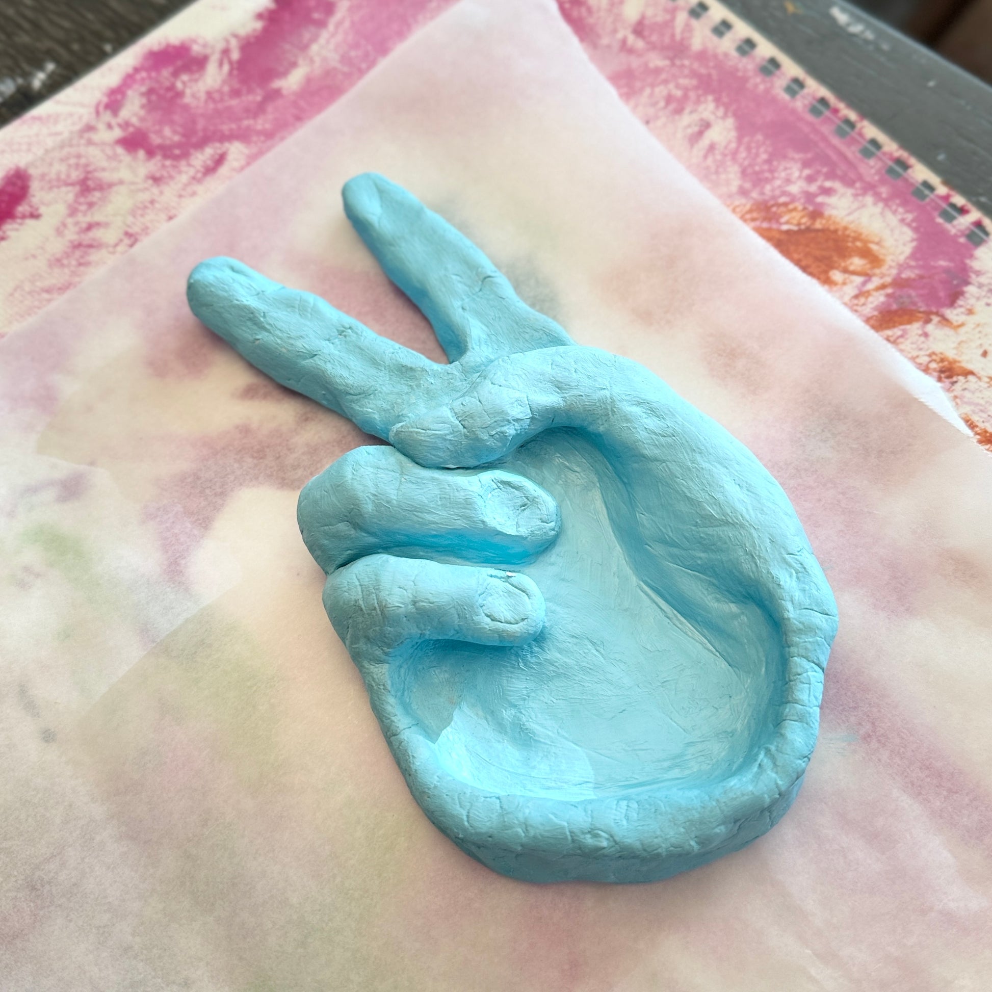 Clay scultpted hand holding a peace sign on a white and pink painted surface