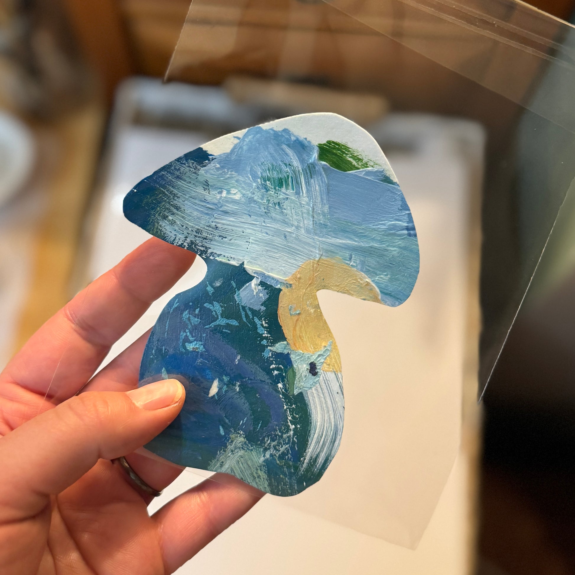 The artist holds a thick, large, blue handmade sticker. The sticker is a mushroom with a thick base and a wide mushroom cap. Details of the color palette come through in the handmade art sticker, crafted from the artist’s paint palette.