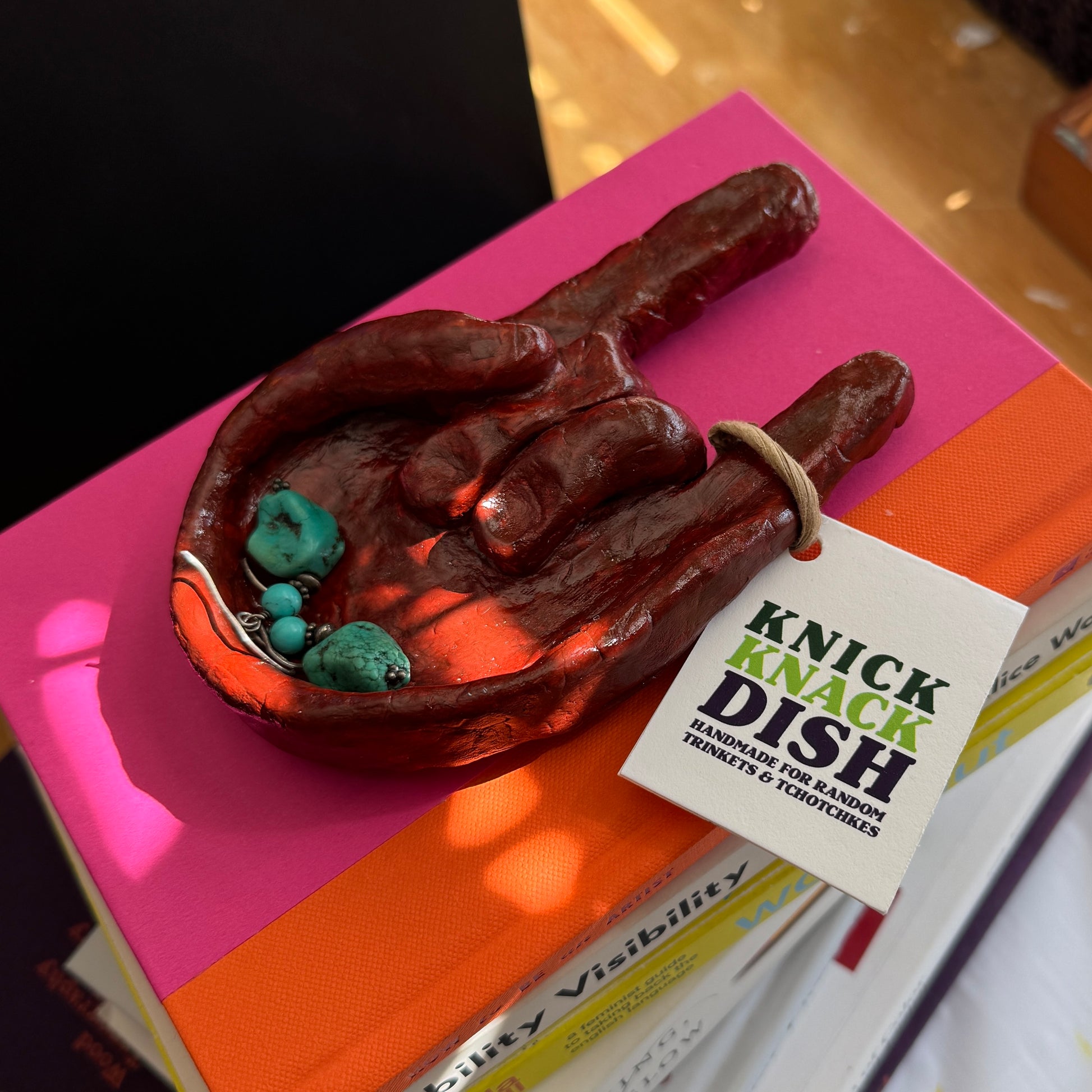 Decorative dish shaped like a hand holding teal earrings, placed on books with a 'Knick Knack Dish' tag.