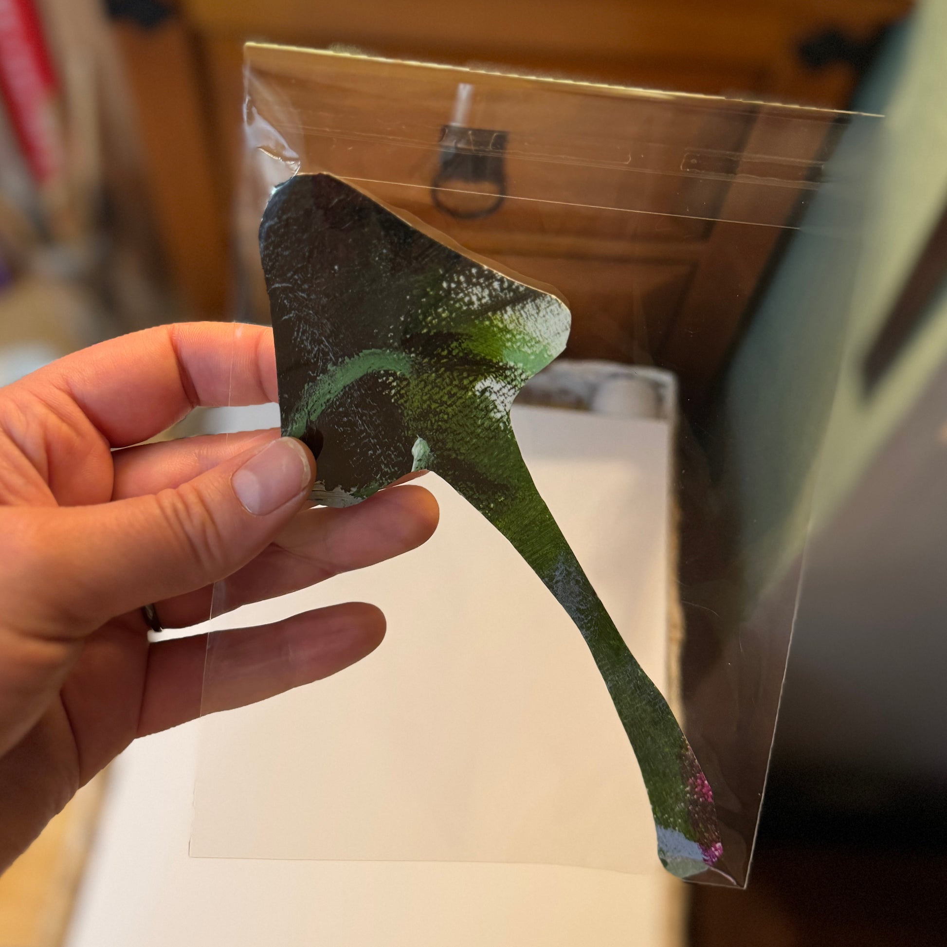 Large art mushroom sticker created from the artist’s paint palette. The sticker has a triangular mushroom cap and a long, thin green stem. The artist holds the sticker in their studio next to a wooden cabinet.
