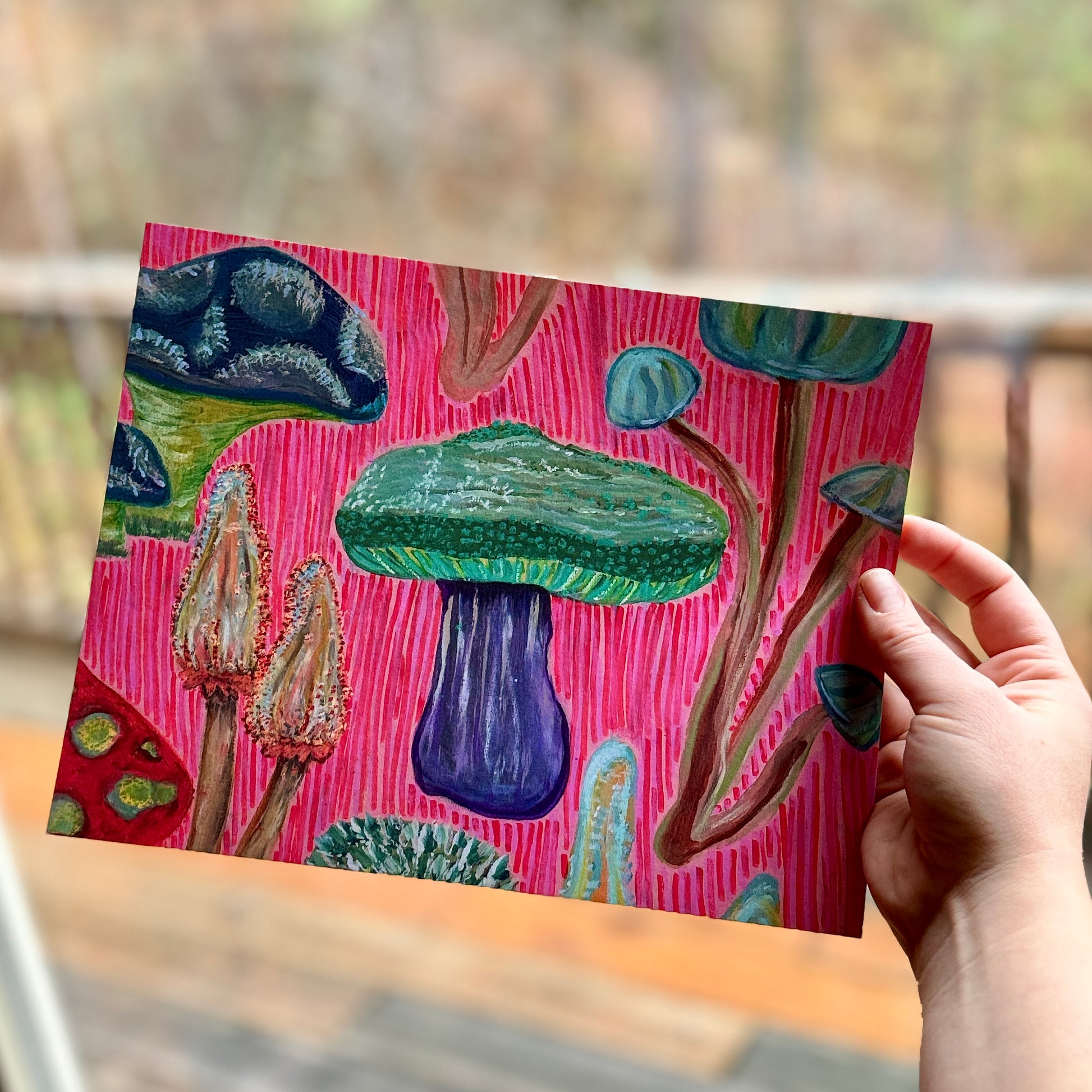 Small, abstract fine art print, Fun Fungi, held by the artist in a cabin window, facing nature. Abstract artwork features whimsical mushrooms in vibrant colors, combining playful design with organic textures.