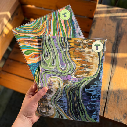 Artist holds two abstract prints, Flowing Through Earth and The Never-Ending Spiral. Prints are vibrant, colorful, and shining in the natural light. 