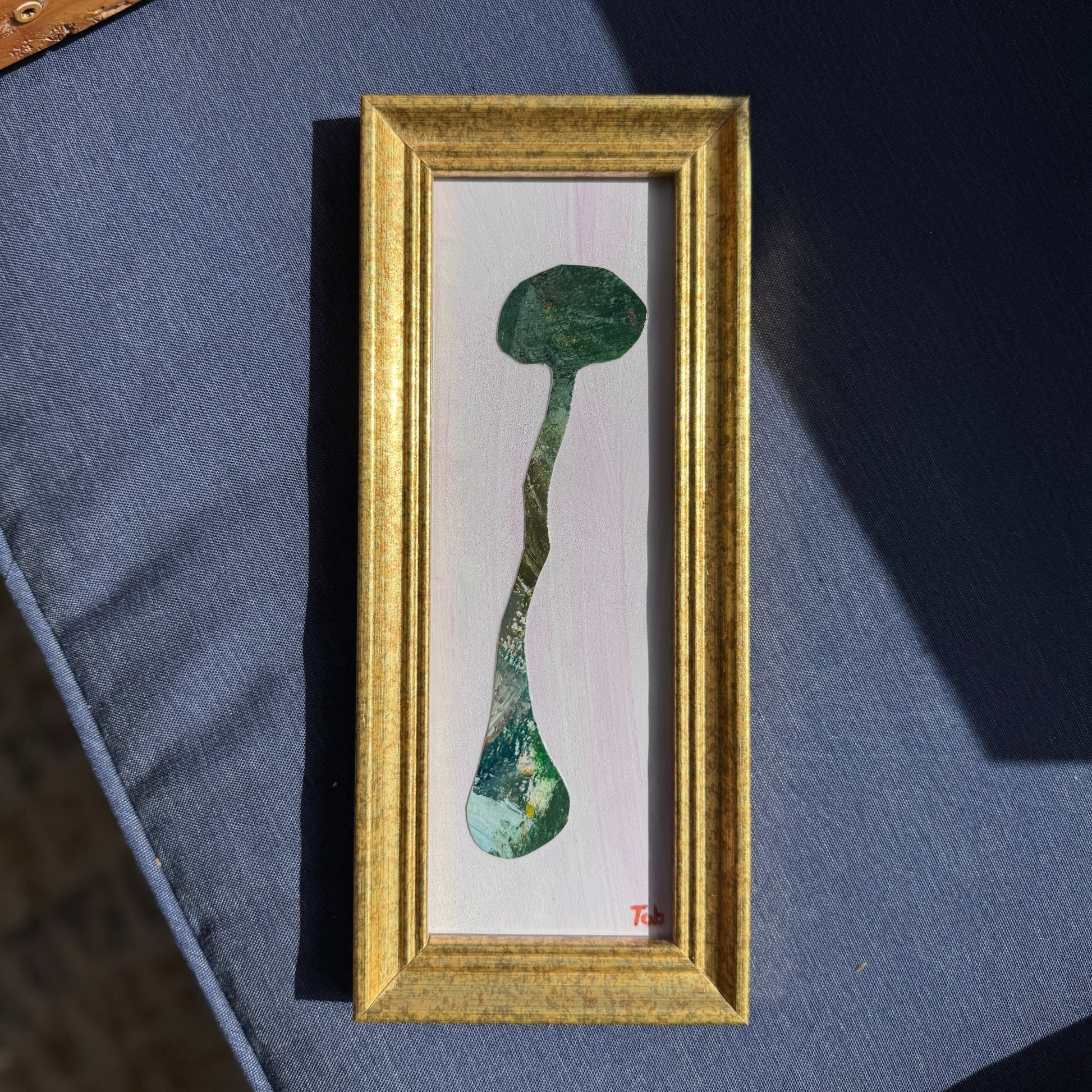 Framed artwork of a long green paper collaged mushroom on a blue surface.