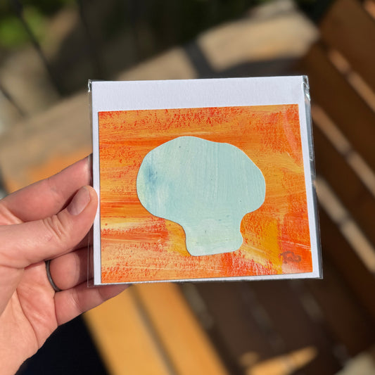 Hand holding a small collage artwork with a mushroom design on an orange background.