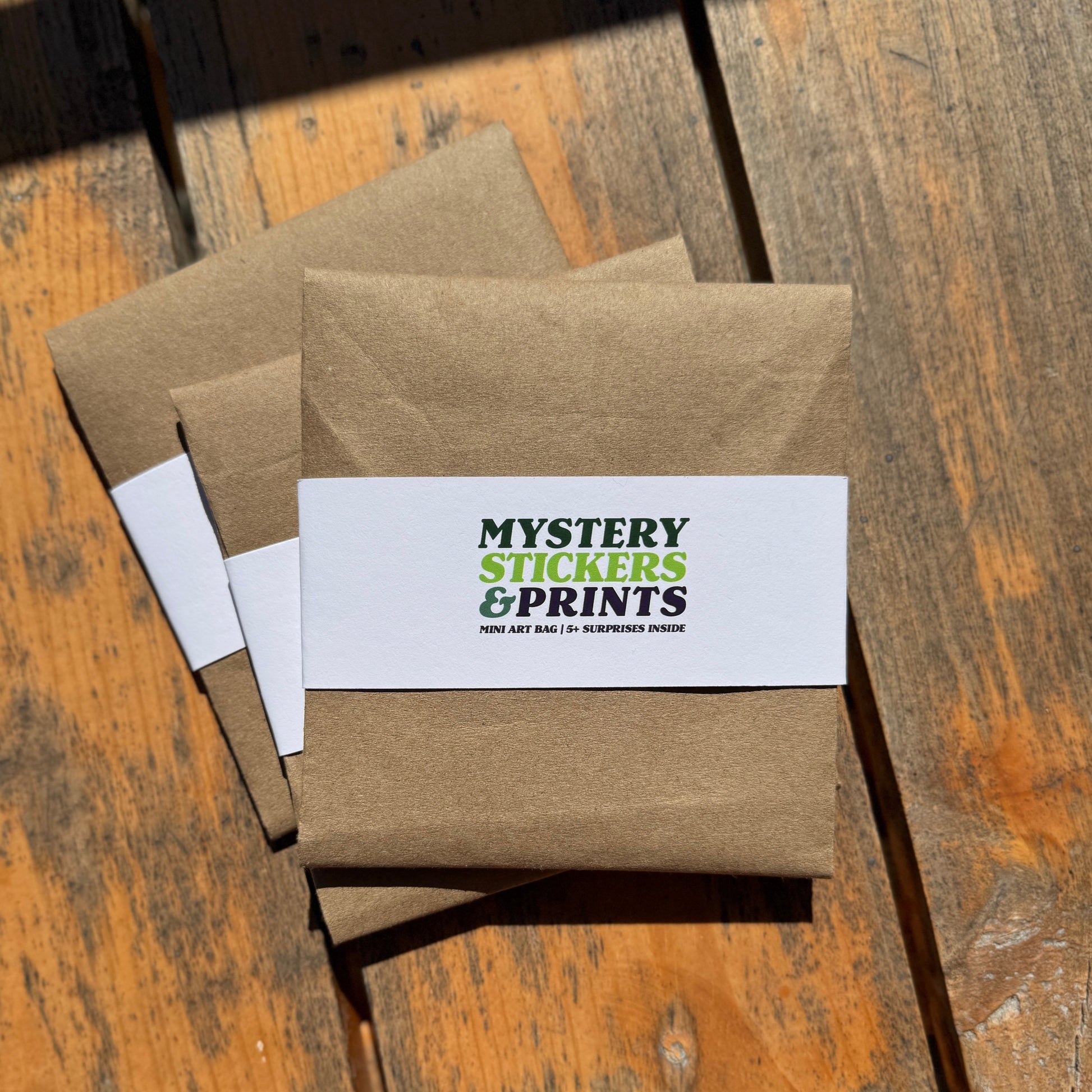 Brown paper packages on a wooden surface with 'Mystery Stickers and Prints' label.
