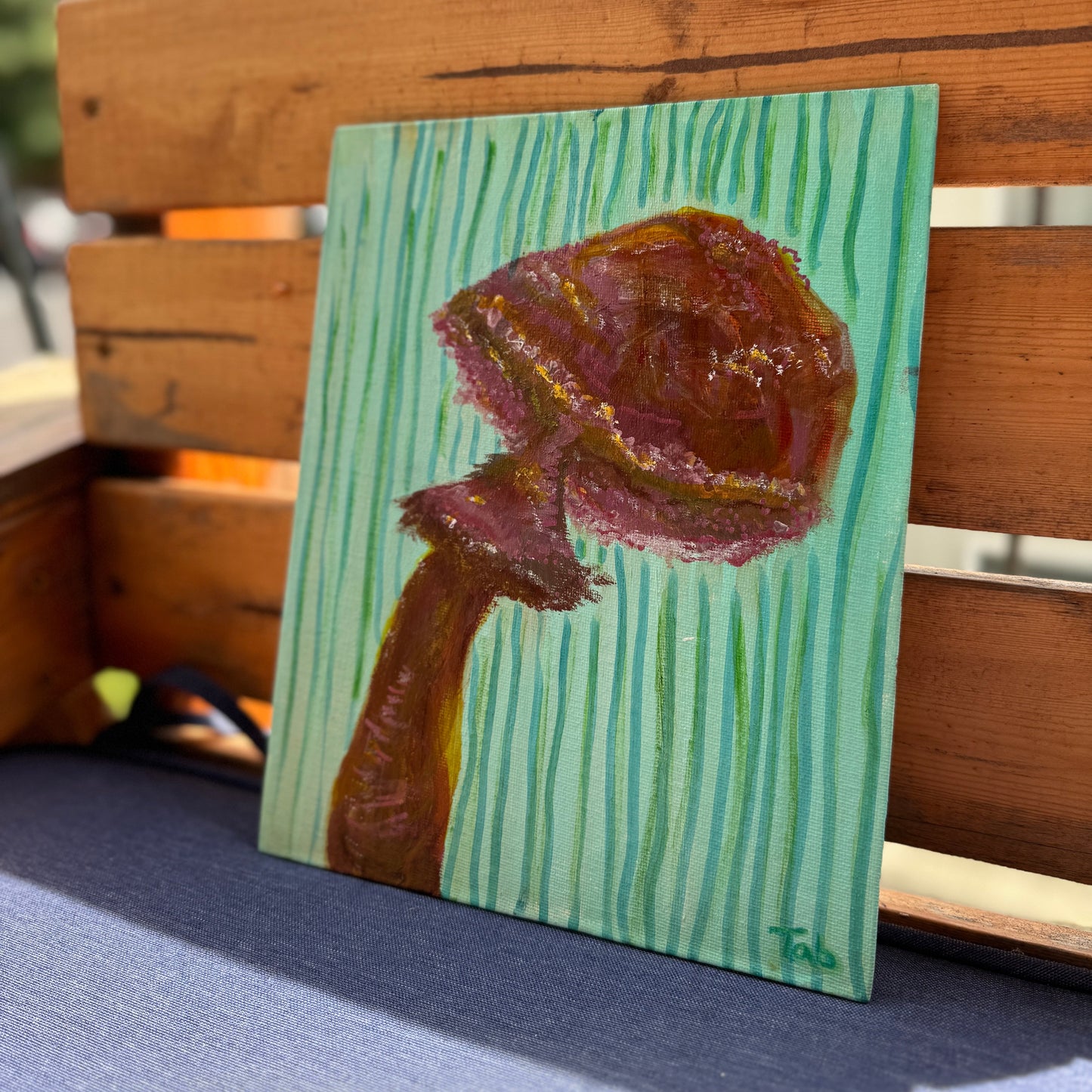 Small painting of a brown mushroom on a green striped background, leaning against a wooden surface.