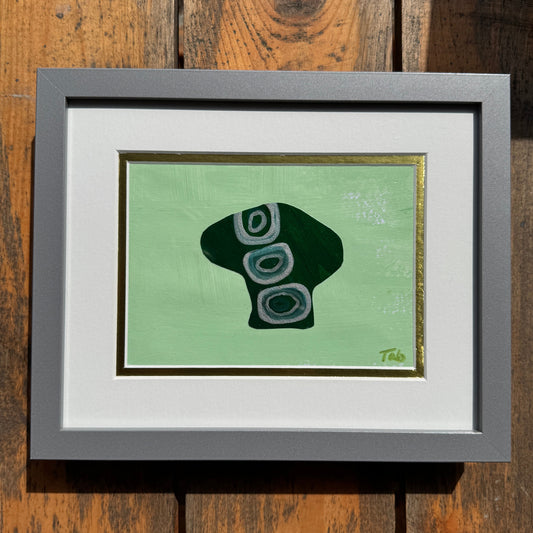 Small green mushroom collage artwork in silver frame on a wood background.