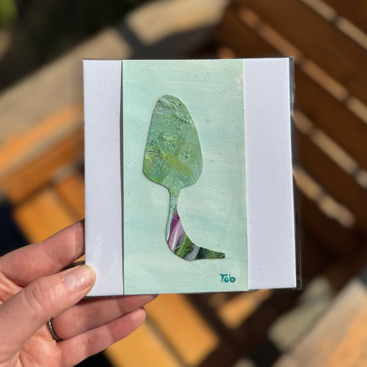 Hand holding a small teal artwork depicting an abstract mushroom collage.